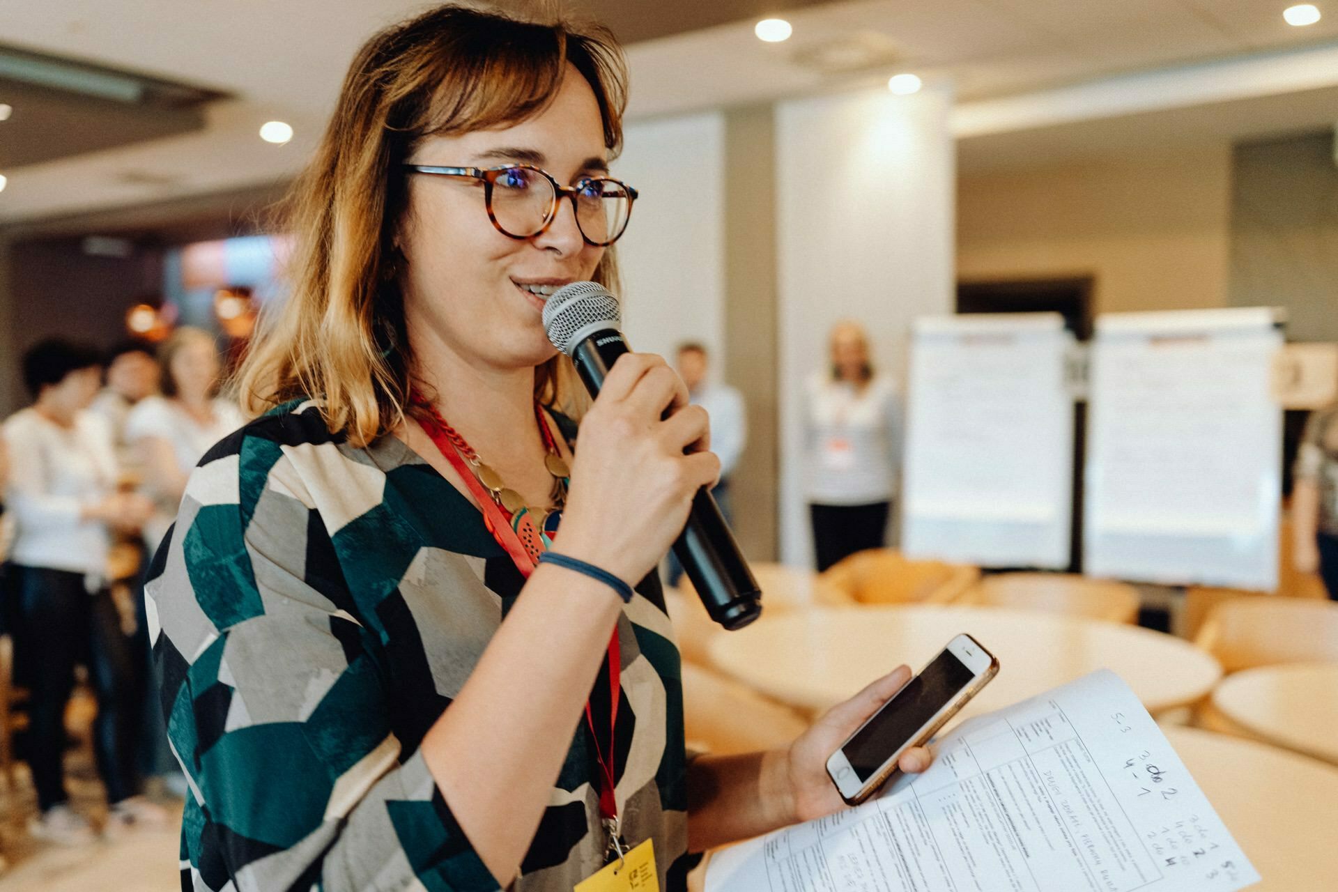 A woman wearing glasses and a patterned shirt holds a microphone and speaks at an event held in a room. In her other hand she holds documents and a smartphone. In the background, people are gathering, and boards of notes can be seen, captured by a photographer from Warsaw providing photo coverage of the event.  