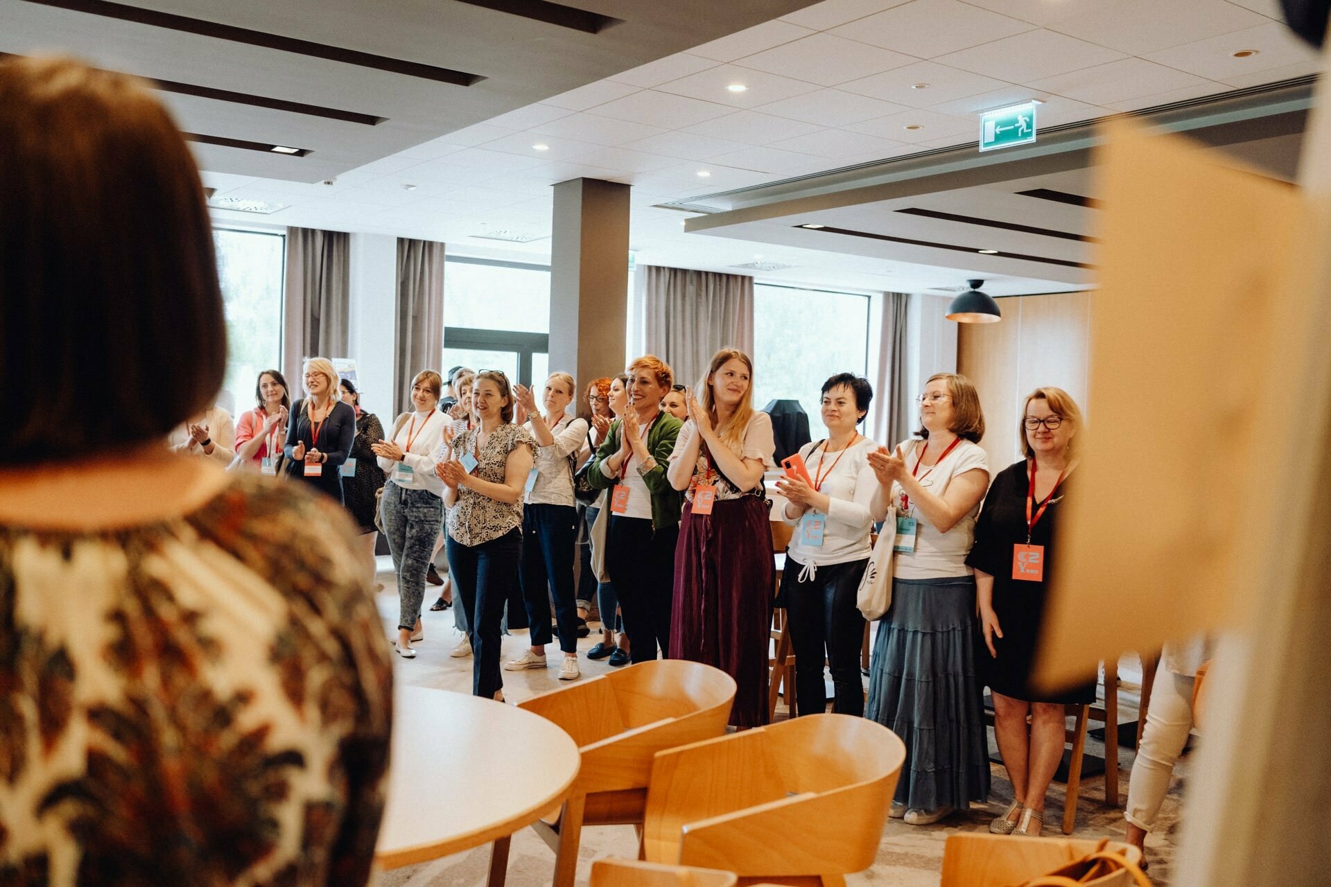 A group of people, mostly women, are standing in a well-lit room with large windows. They clap and seem engaged and attentive. Some are wearing name badges, and the atmosphere seems positive and supportive. In the foreground are light wood chairs and tables, perfectly captured by event photographer Warsaw.   