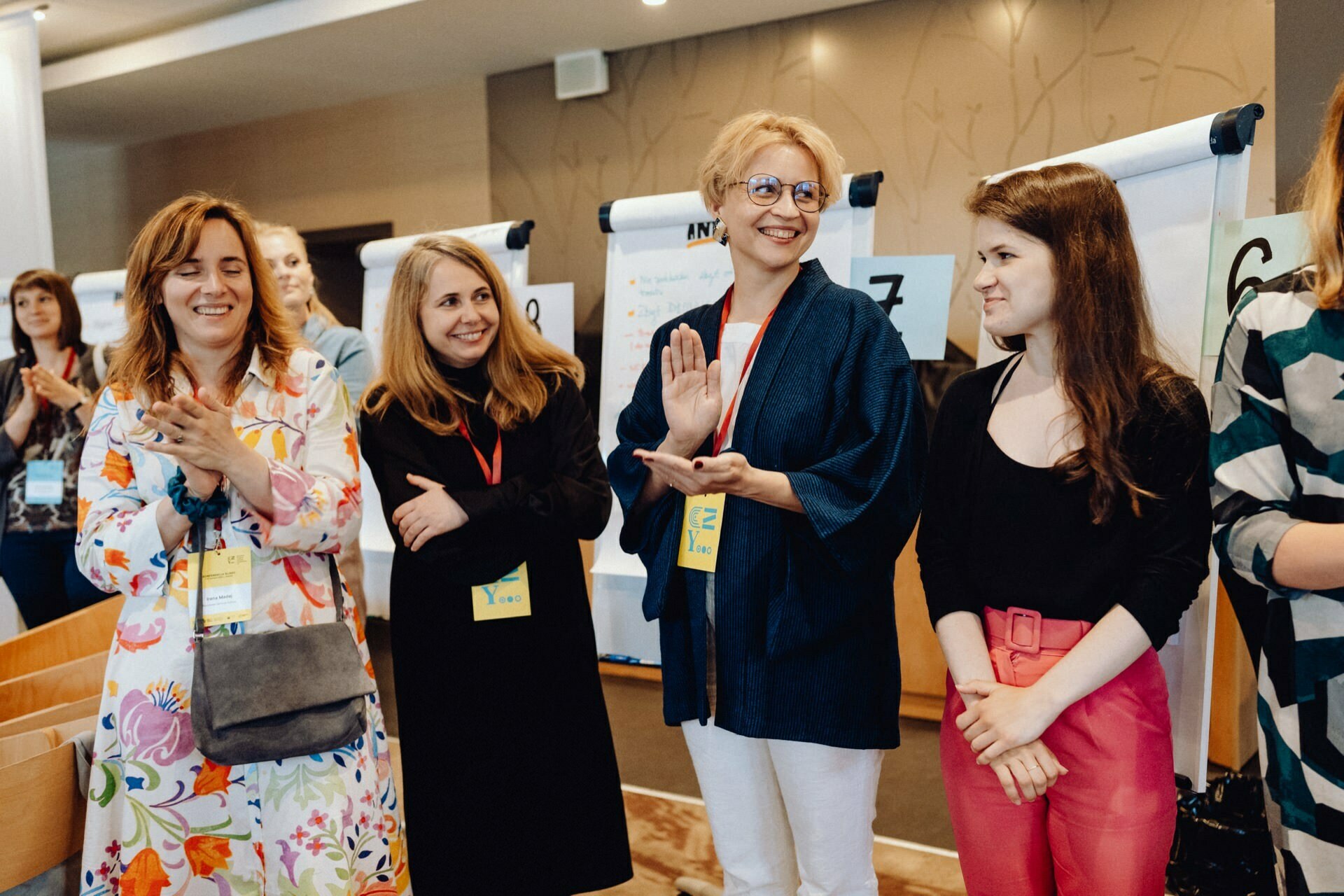 A group of five women standing in a room, smiling and carrying on a conversation. They sit in front of white flip charts, each wearing a name badge around their neck. The background is light brown walls with a subtle leaf pattern. This captivating photo essay of the event shows their lively interaction.   