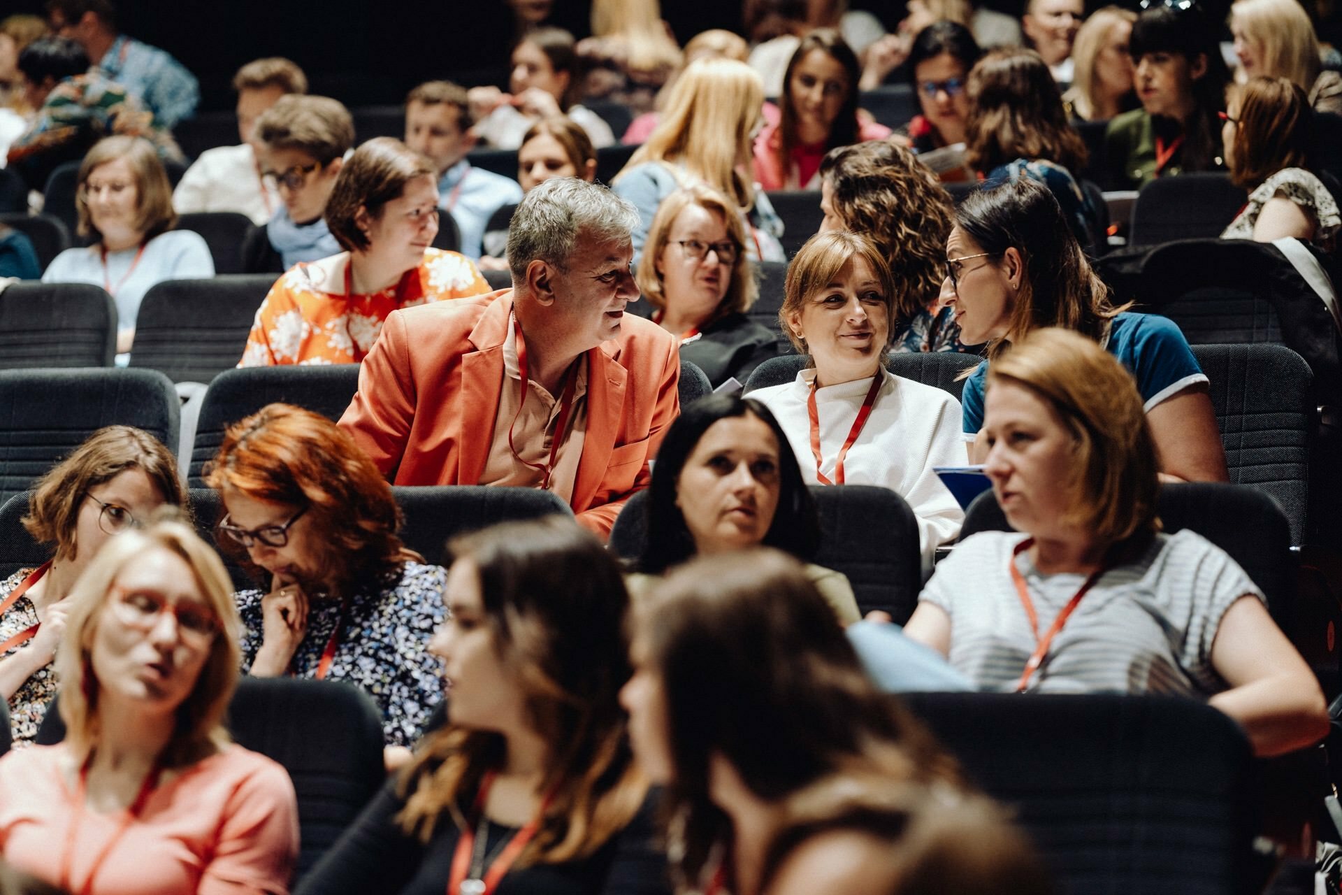 A diverse group of people sitting in the auditorium are engaged in conversation before the event begins. Some are wearing lanyards and their faces vary, indicating engaging discussions. In the background, you can see rows of chairs filled with attendees, perfectly capturing the atmosphere of the photojournalism of the event.  