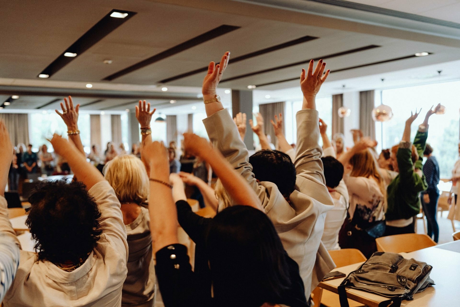 A group of people in a spacious, well-lit room enthusiastically raise their hands. They are seated on chairs arranged in rows, and the blurry background suggests a communal or celebratory event perfectly captured by an event photographer Warsaw. 