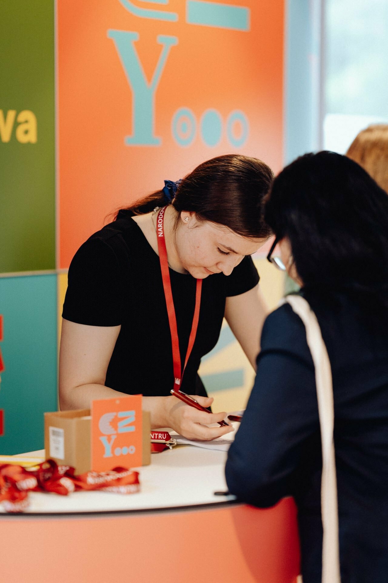 A woman in a black shirt and red lanyard helps another person at a colorful booth. The booth, captured in a photo essay from the event, has vibrant panels in orange, blue and green, with text partially visible. The tabletop features an open box and more lanyards.  