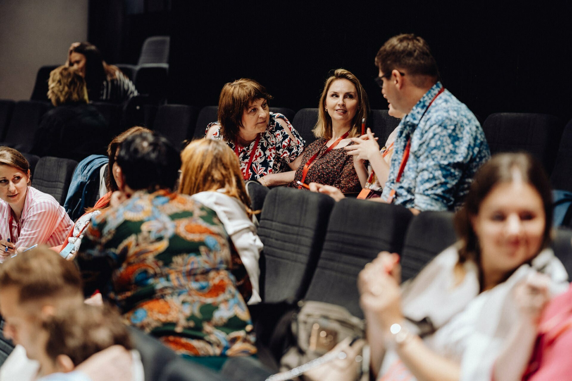 A group of people sitting in theater-style chairs, immersed in conversation. Some wear badges on lanyards around their necks. A woman in the middle smiles, listening to a man talking. In the background you can see more people sitting and talking, beautifully captured by a photographer from Warsaw.   