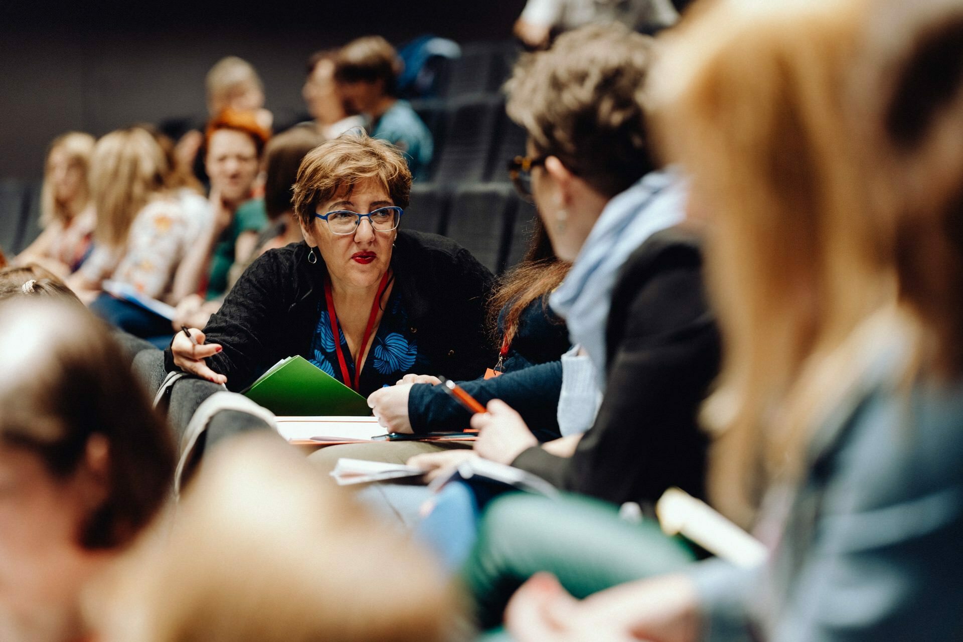 A group of people are sitting in an auditorium having a discussion. The focus is on a woman with short hair and glasses, holding a green folder and talking to the person next to her. The auditorium seems to be busy with other attendees talking and taking notes, capturing the essence of this dynamic photo essay of the event.  