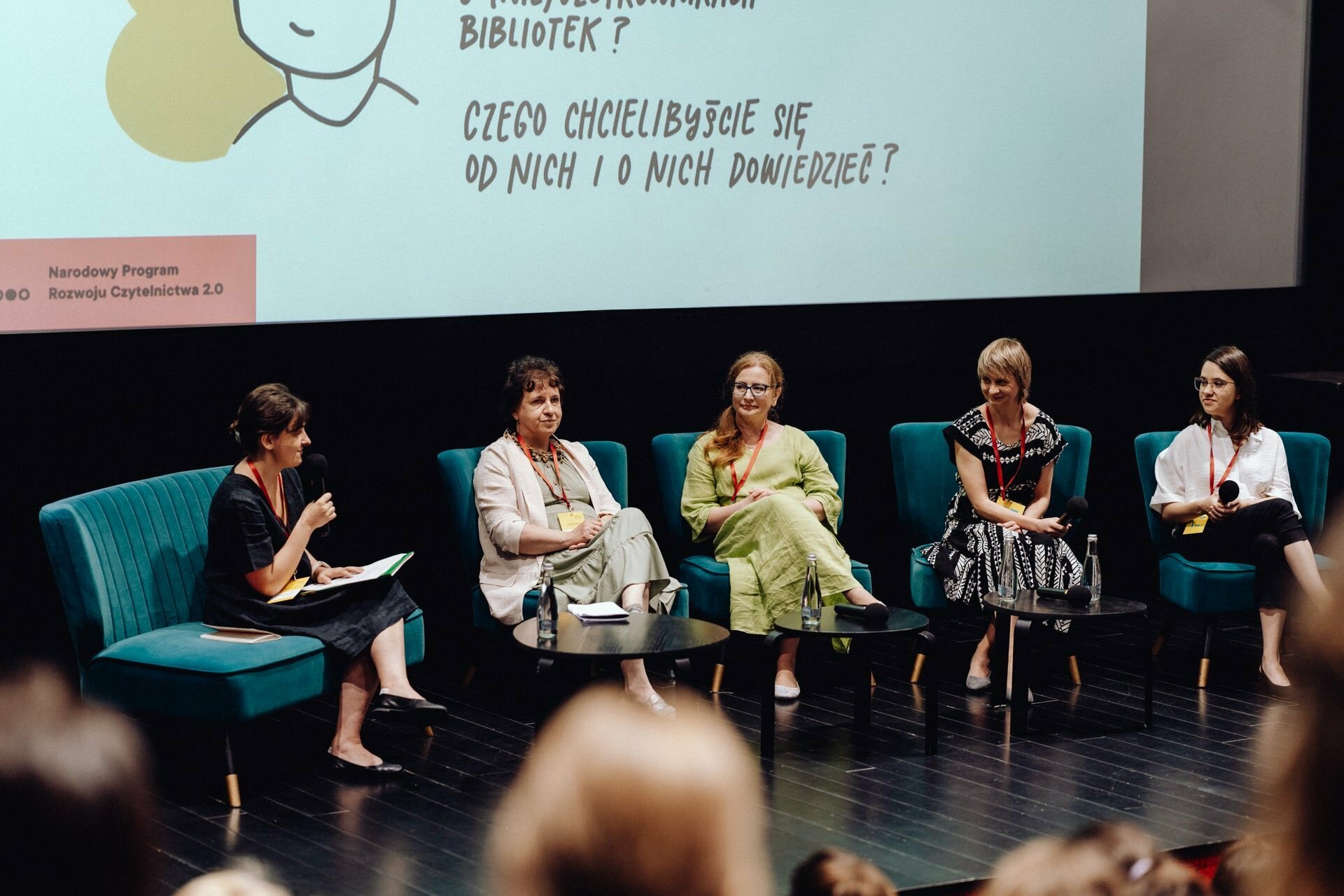 Five people participate in a panel discussion on a stage with a background displaying text and illustrations. They sit on turquoise chairs and a black couch. One person speaks into a microphone while holding notes. The others listen carefully to what the photographer at the event has captured for the event photo essay.   