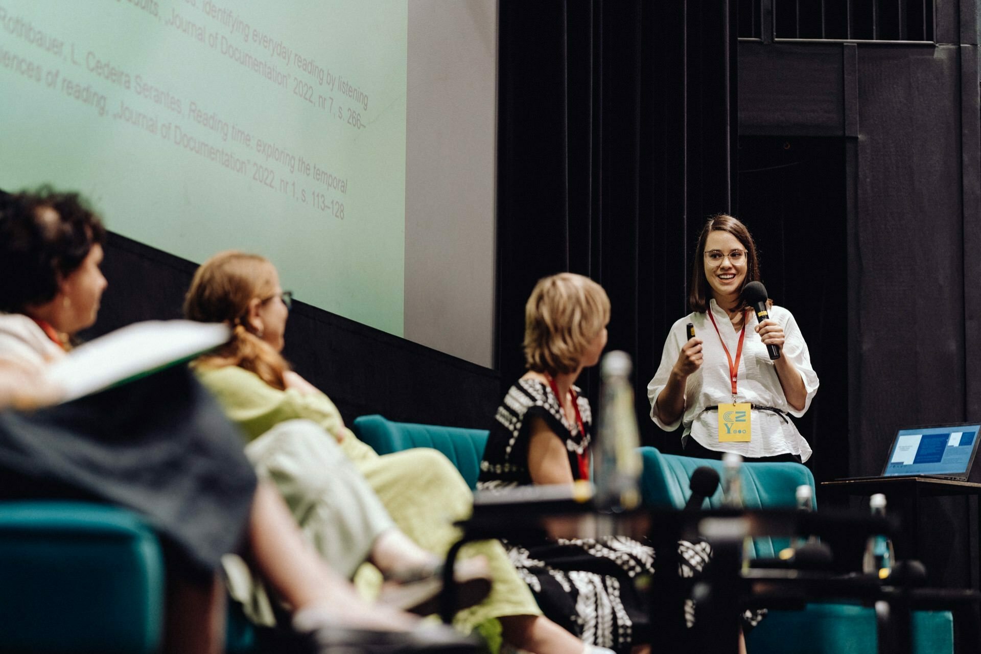 Four people sit in a panel on the stage, while another person stands and speaks into a microphone. In the background is a large screen displaying text, and a laptop is visible on a small table. The atmosphere is either academic or professional - a perfect photo-reportage of the event captured by a photographer from Warsaw.  