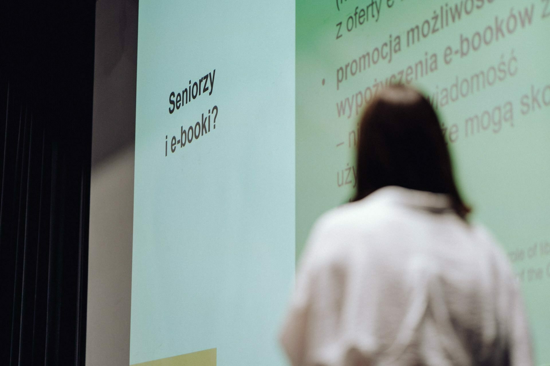 A person standing in front of a large screen and giving a presentation. The screen displays text in Polish, with the main visible text "Seniors and e-books?" A professional event photographer from Warsaw captures the moment, taking care to blur the background to emphasize the content of the presentation.