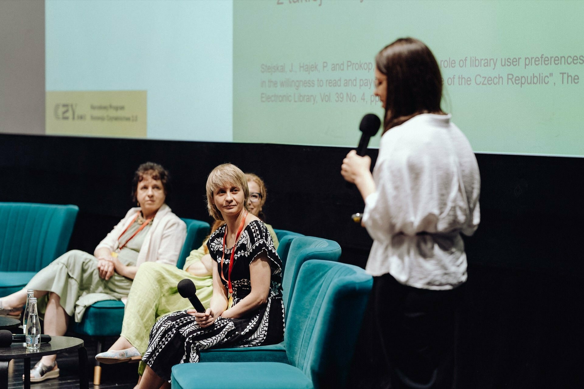 Four people sit on turquoise chairs on the stage and lead the discussion. The person standing to the right holds the microphone and addresses the seated panelists. As a presentation slide is projected on the screen in the background, every moment of this engaging session is captured by a photographer at the event.  