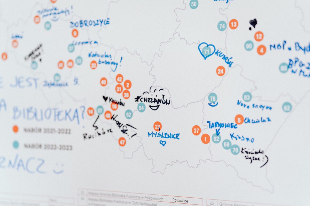 A map board filled with a variety of colorful markers, symbols and handwritten notes from a photo recap of the event. Blue and orange circles indicate various locations, numbers and labels. The handwriting includes place names and scribbles, including a smiley face and a heart in the Myslenice area.  