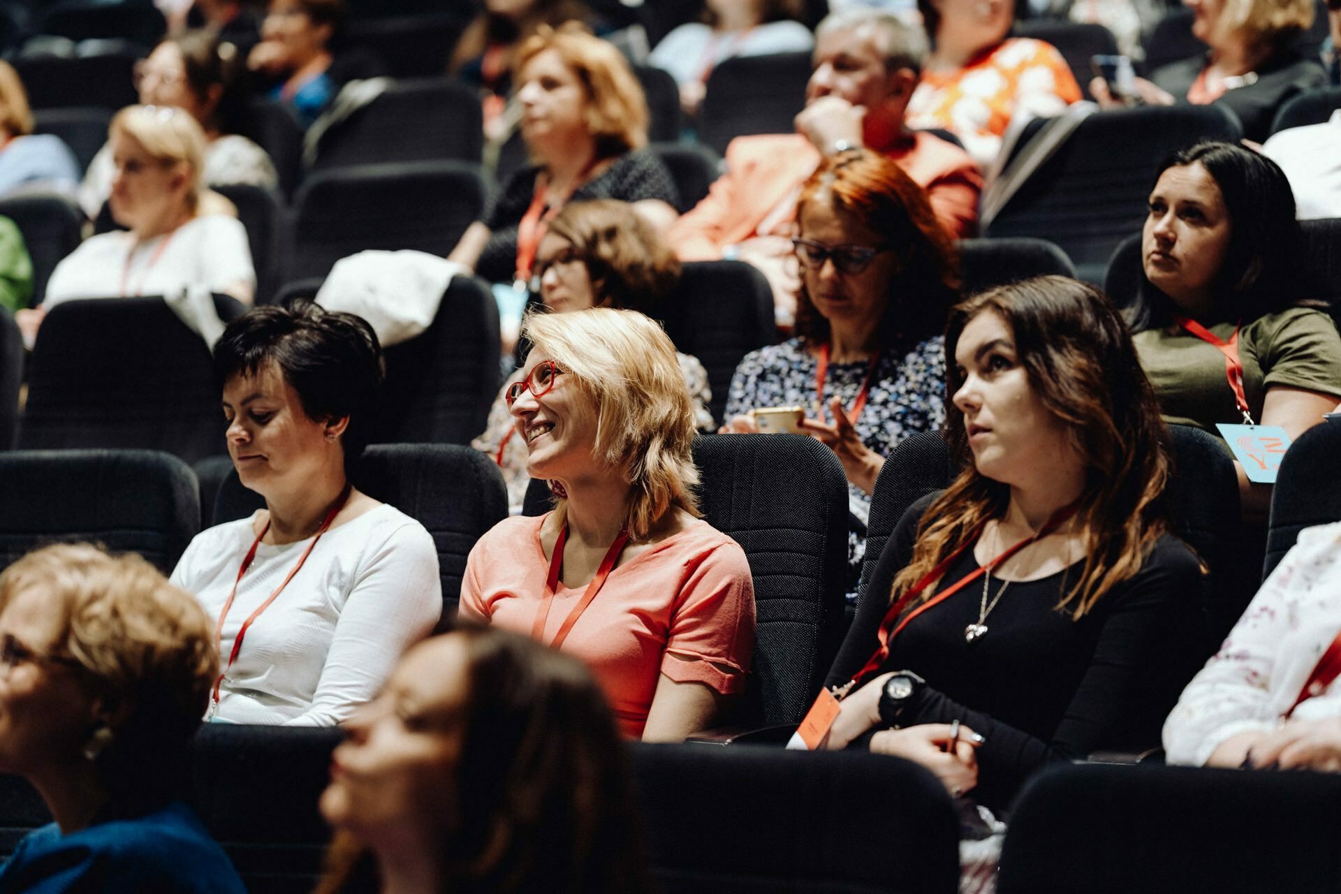 A group of people, mostly women, sit in rows of seats at an event or conference. Many are wearing attendee badges and a few are taking notes. The atmosphere seems focused and engaged, which was perfectly captured by the photographer for the event, who provided a detailed photo report of the event.  