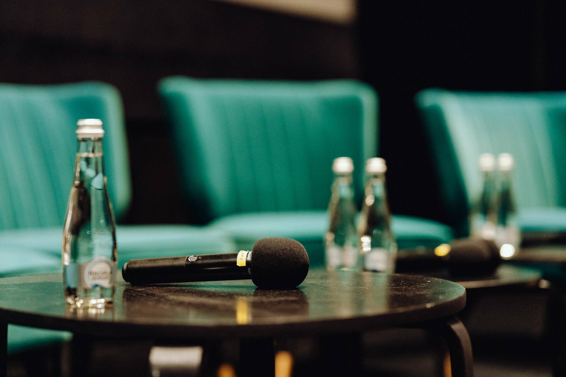 A black microphone sits on a round table surrounded by several clear glass water bottles. Three turquoise-colored chairs are set up in the background, suggesting a setting for a panel discussion or interview, all perfectly captured by the photographer at the event. 