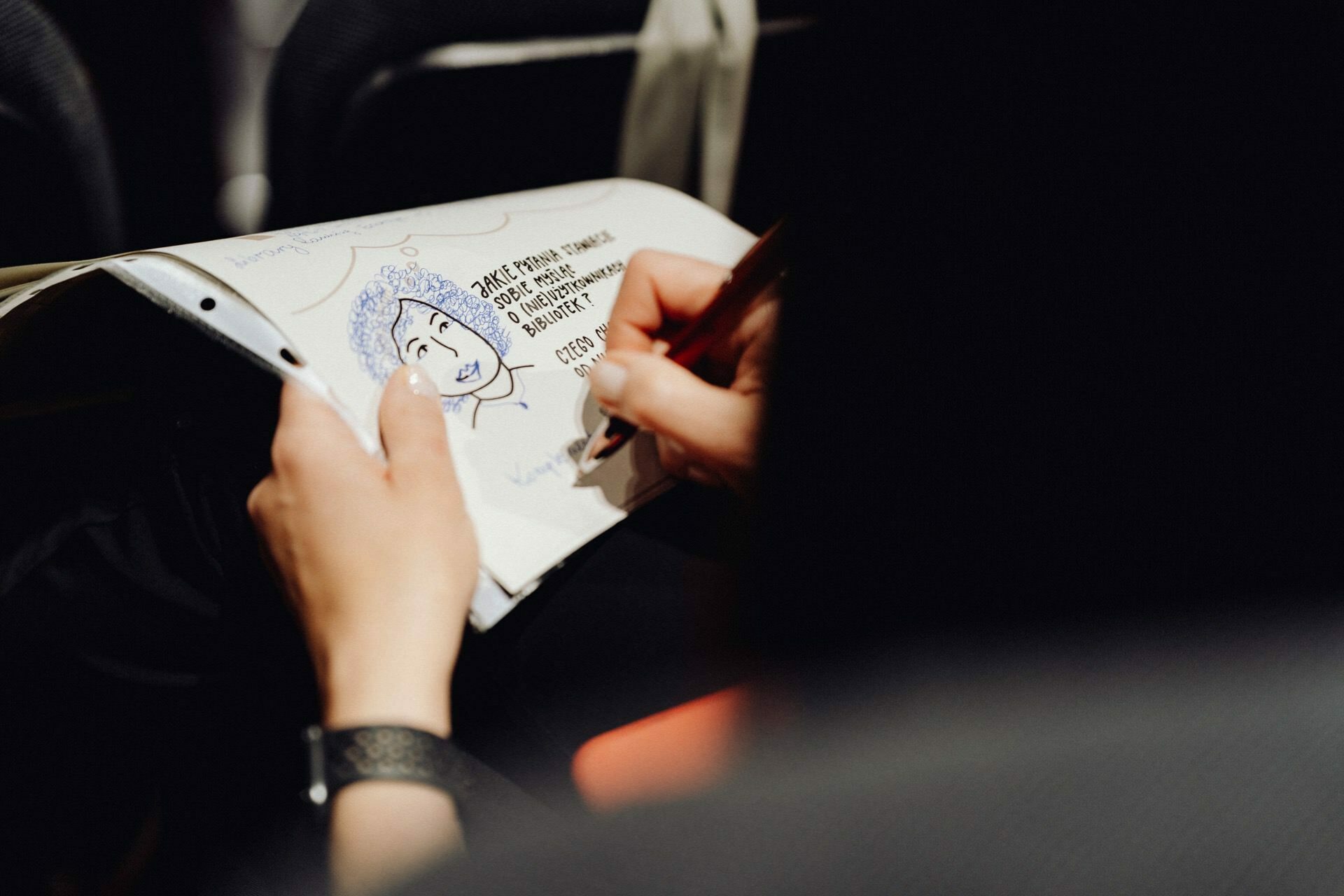 In a public place, such as a conference or classroom, someone sits and draws with a pen in a sketchbook. The sketch includes a cartoon face and some text. The background is out of focus, giving it the effect of an immersive photo essay of the event.  