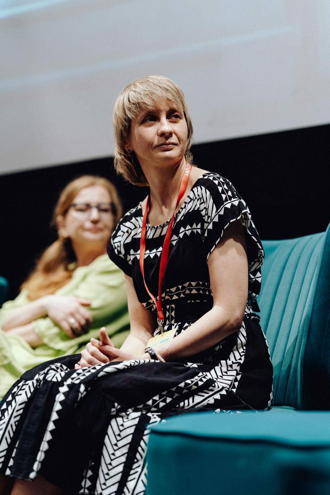 Two women sit on turquoise chairs during a panel discussion. The woman in the foreground, captured by the photographer at the event, is wearing a black and white patterned dress and a red bow, looking slightly to the side. The woman in the background is wearing a light green outfit and glasses.  