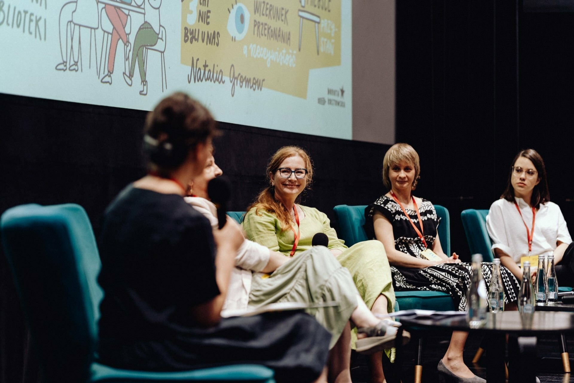 A panel of five women sit on turquoise chairs on a stage and lead a discussion. One speaker speaks, the others listen attentively. Behind them, a large screen displays a colorful illustration and text. They all wear red lanyards around their necks - which was perfectly captured by a photographer at the Warsaw event.   