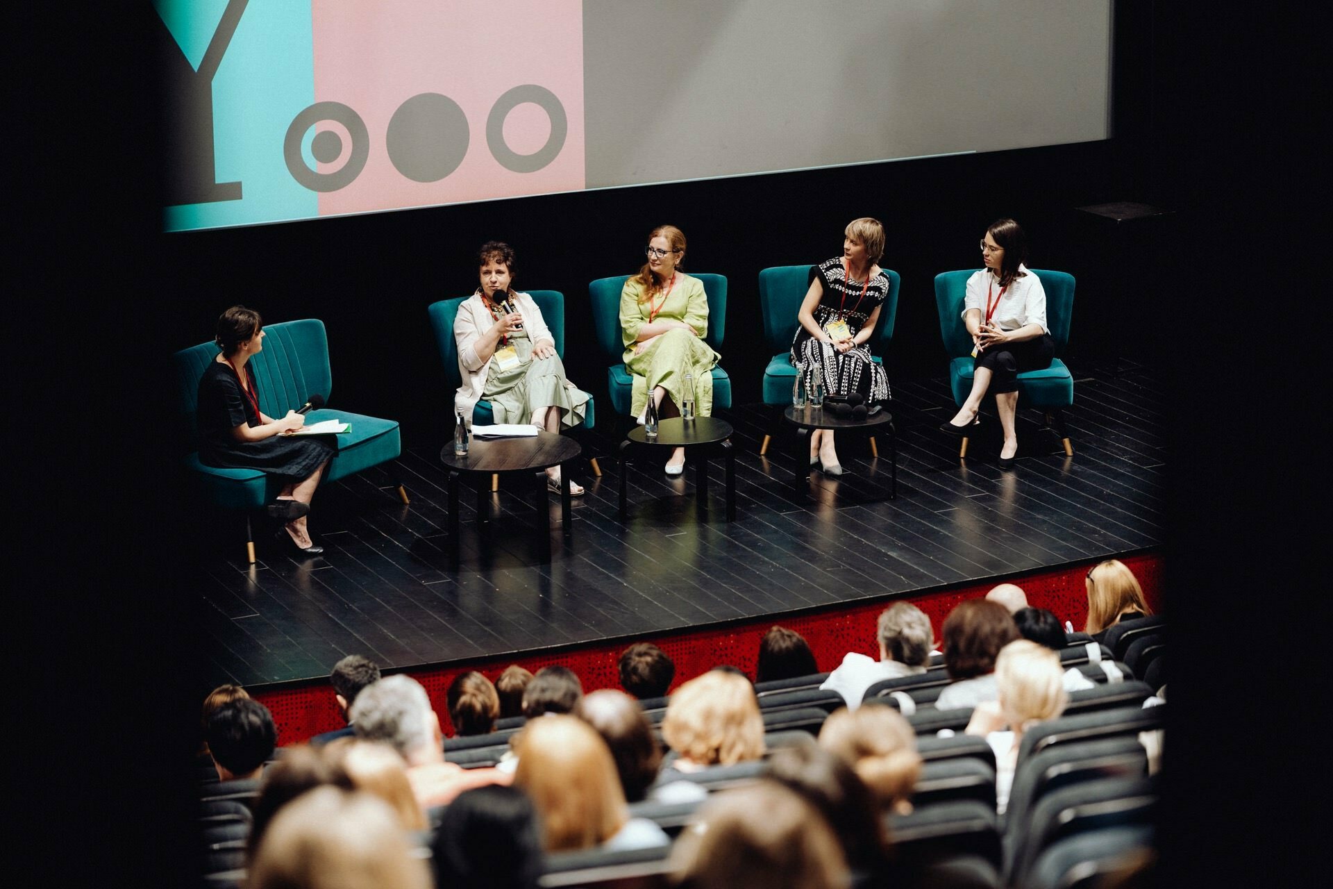 A group of five people sit on stage during a panel discussion, each with a microphone. Four sit in chairs, one speaks; the fifth person sits at a small table. The audience faces them, while a color screen glows in the background - perfect for photo coverage of the event by any talented photographer from Warsaw.  