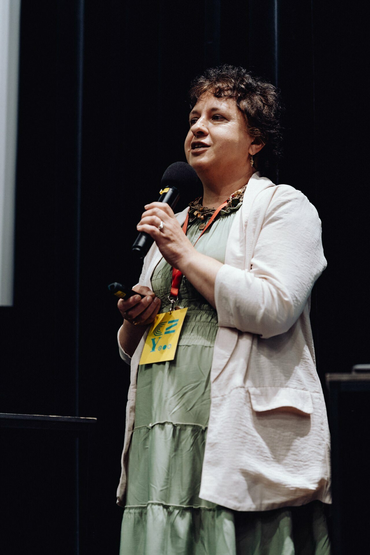 A person with curly hair, wearing a green dress and a bright cardigan, speaks into a microphone on stage during the event. They have a yellow name badge hanging from their necks and appear to be giving a presentation, perfect for capturing on any photo event. 