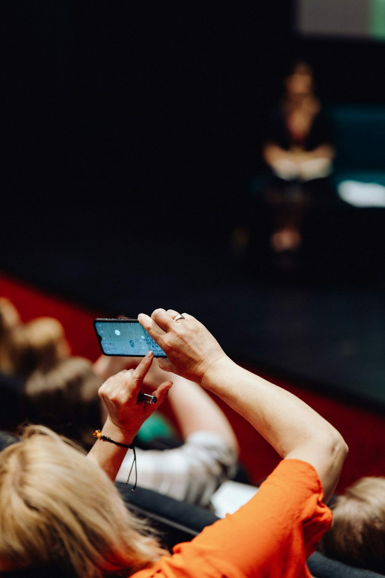 A person in an orange shirt is holding a smartphone up and appears to be taking a photo or video during the event. Blurry figures sit in the background, and there is one person on stage. The atmosphere suggests a seminar or presentation, allowing the person to capture moments that any photographer at a Warsaw event will appreciate.  