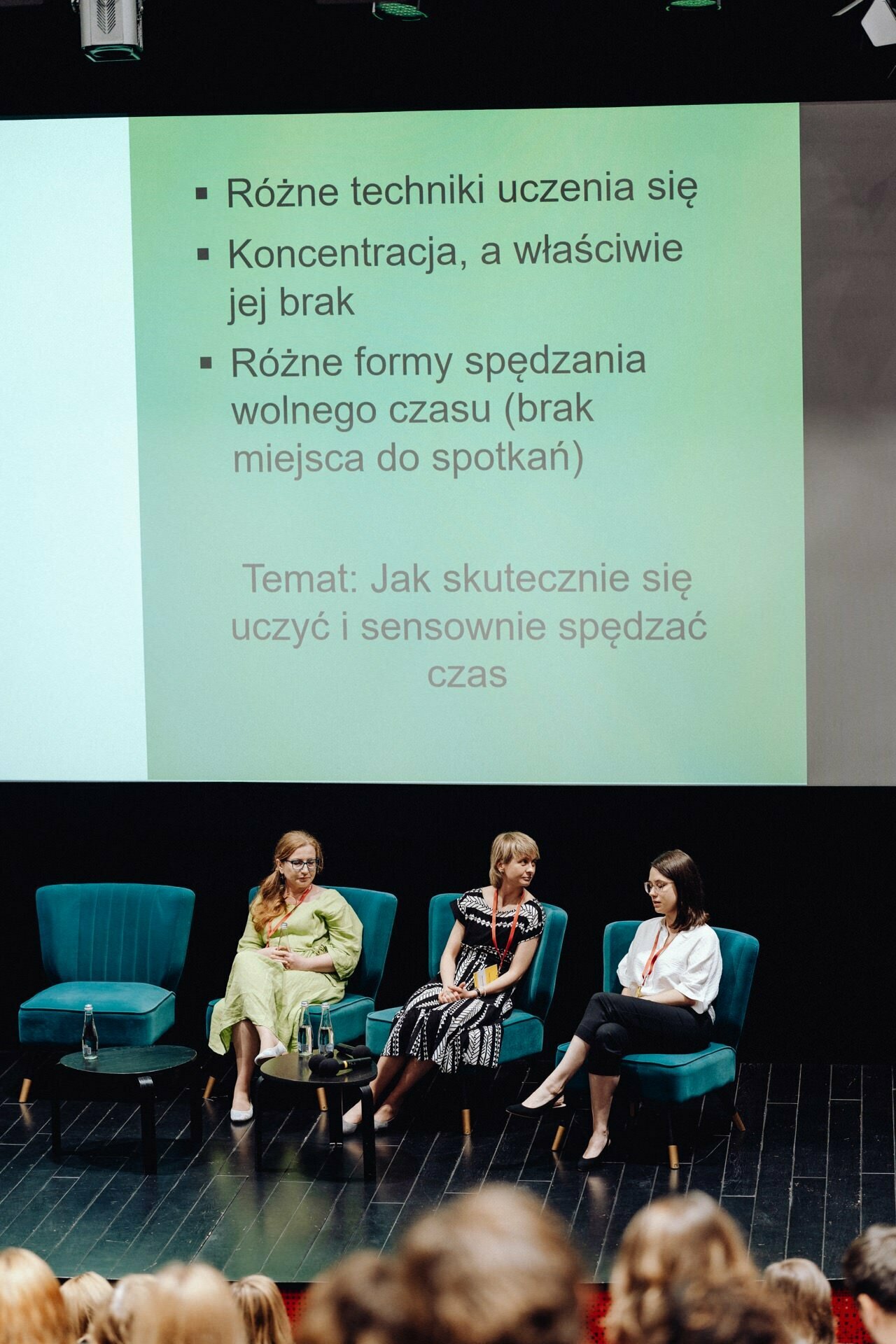 Three women sit on blue chairs on a stage in front of a large screen displaying a presentation in Polish with bulleted points and a highlighted topic. In front of them sits the audience. The text discusses various techniques for learning and spending leisure time effectively, beautifully captured by the photographer at the event.  