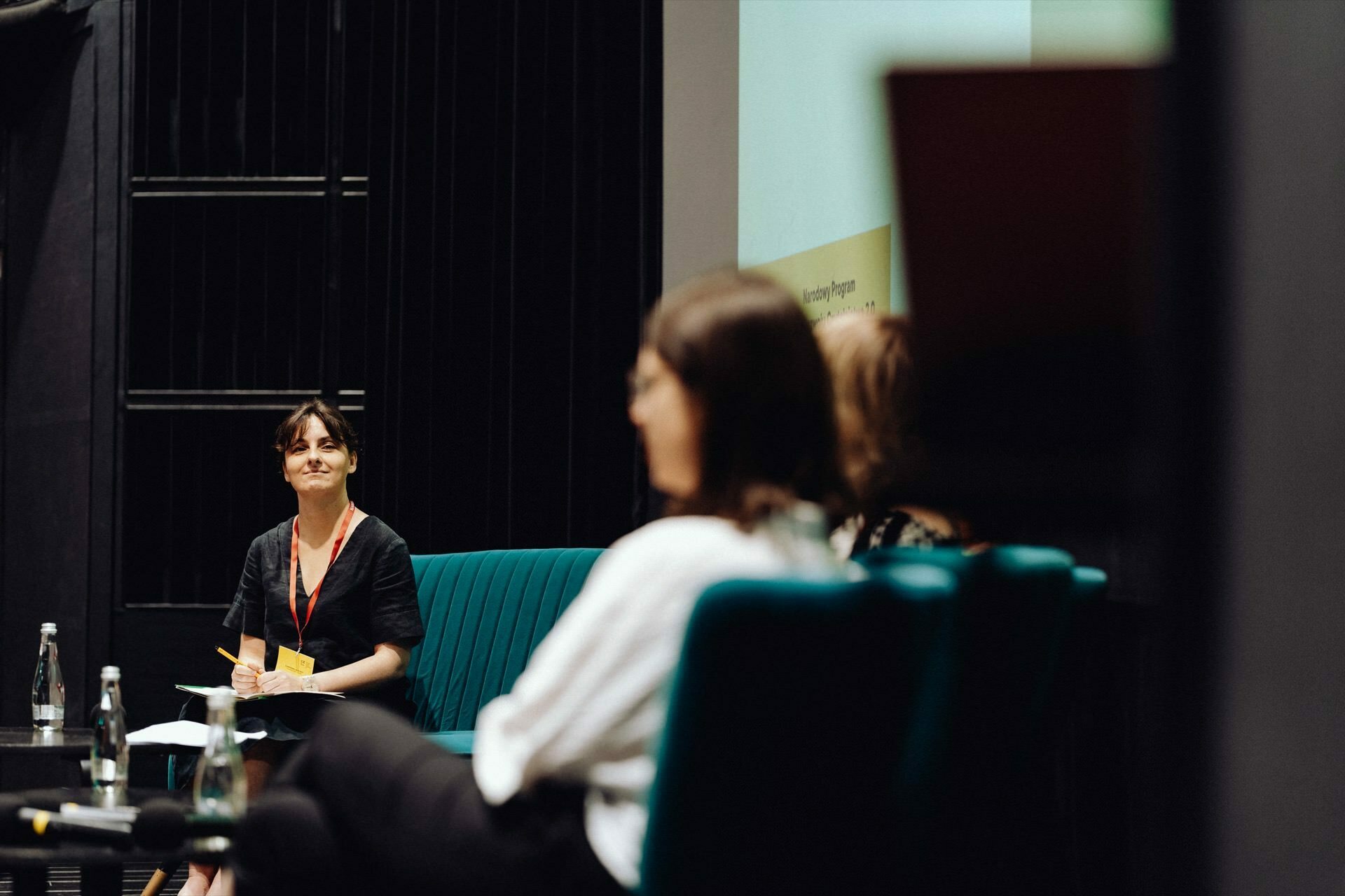 A person sits on stage behind a table with water bottles and papers and takes part in a panel discussion. Two other people sit in the foreground, facing the speaker. In the background is a large screen that captures the essence of this conference or seminar - a perfect photo report of the event by any event photographer.  