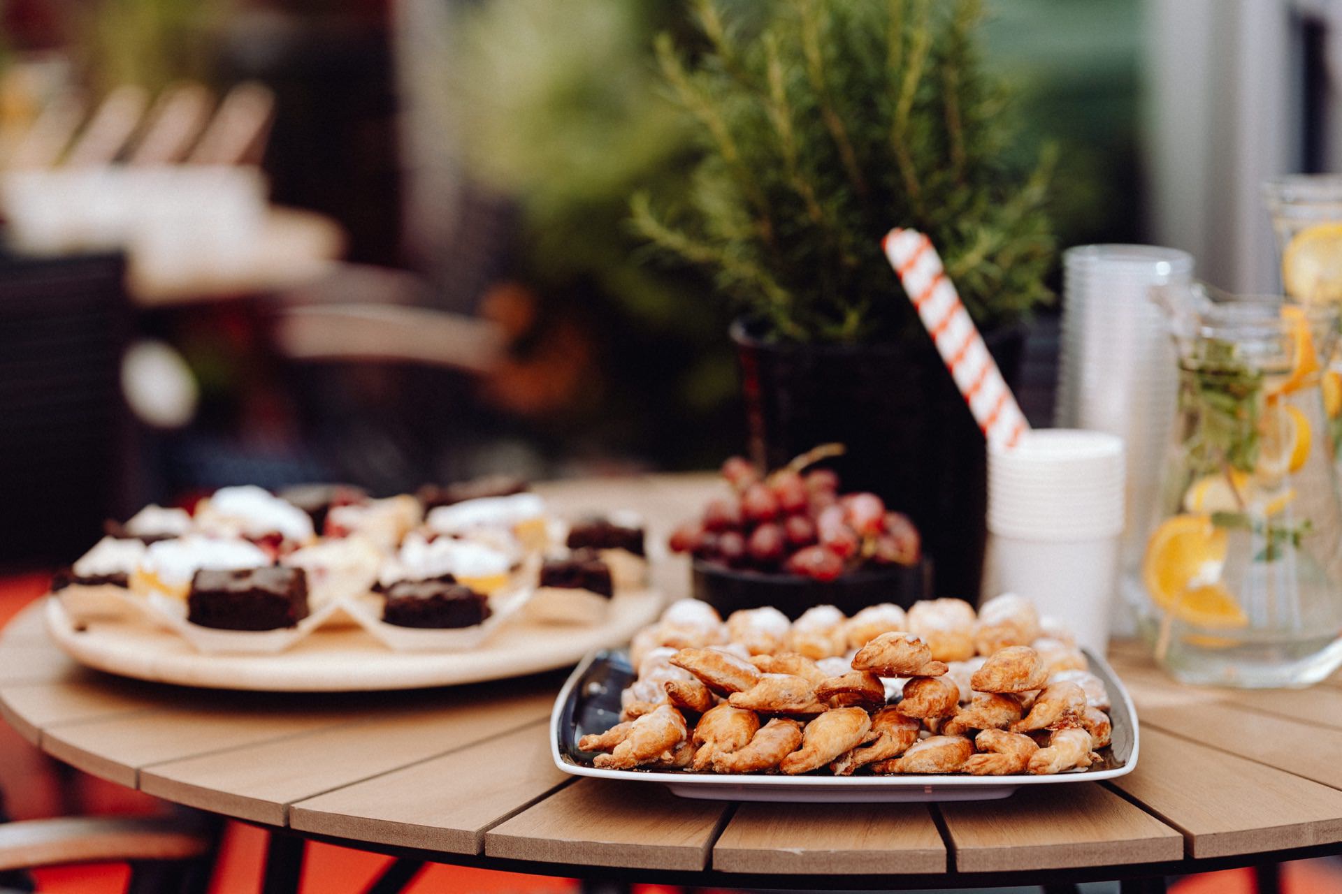 On a wooden table are trays of various cakes, including croissants and cookies, as well as grapes. To the right, cups are stacked next to a jar of lemon-infused water with herbs and a straw. Lush greenery is visible in the background, captured by the photographer at the event.  