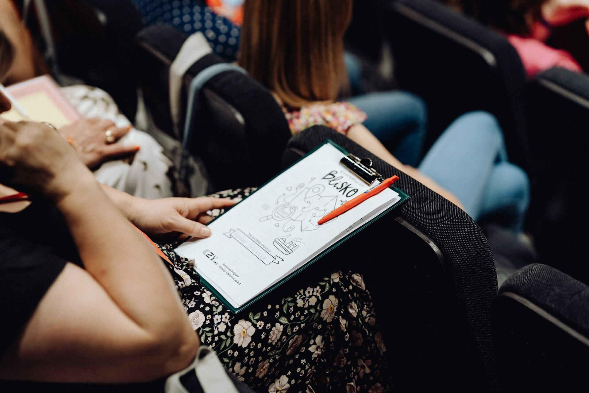 A person sits in the auditorium, holding a pad of paper with a cartoon illustration and the word "PLAYGO" on it. A red pen is attached to the board. The person, who is presumably attending the event as a photographer from Warsaw, is wearing a black top and floral-patterned pants, and other participants are seated nearby.  