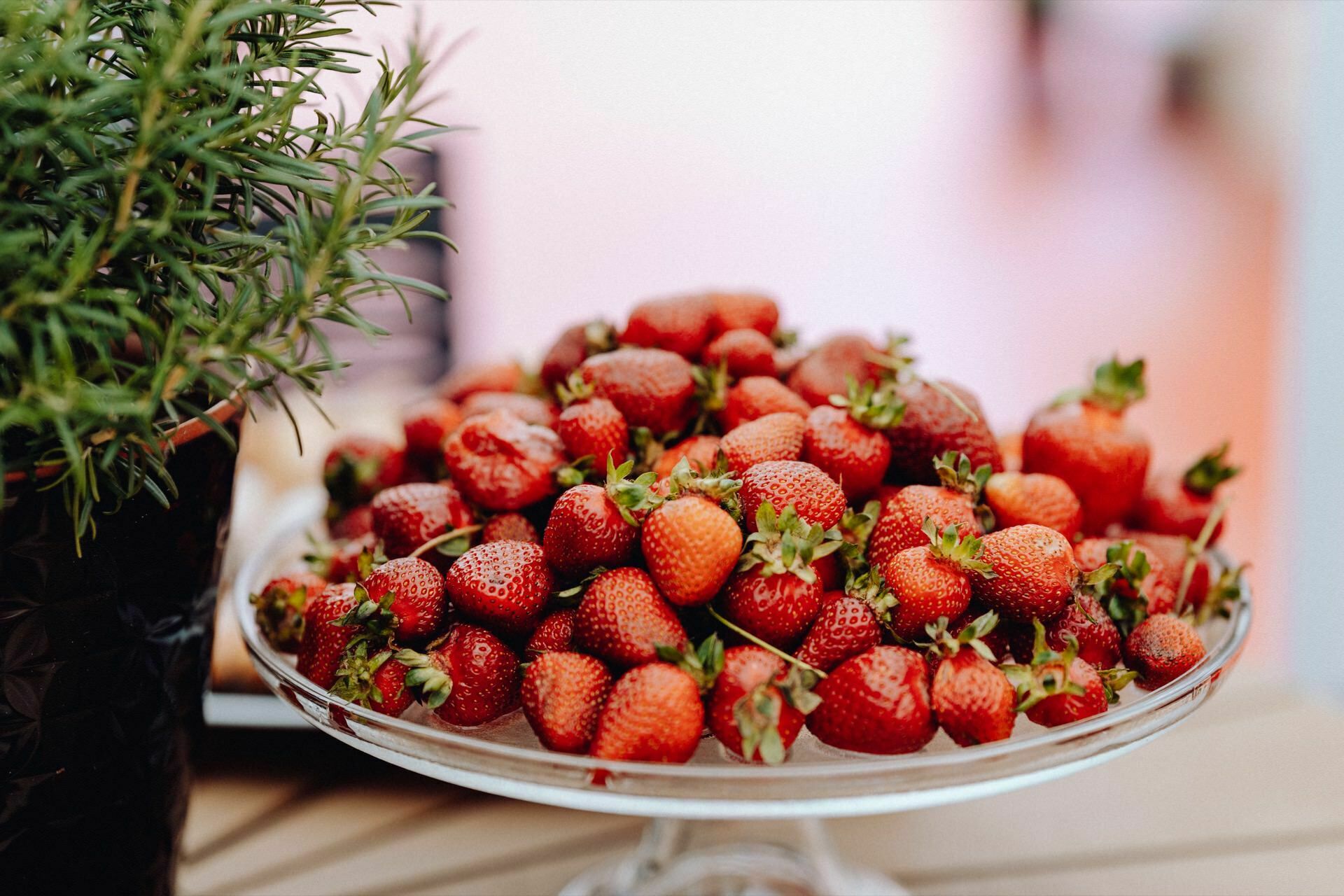 On the table stands a glass plate filled with fresh strawberries. The strawberries are ripe and intensely red, with green leaves still attached. On the left, a sprig of rosemary in a dark pot partially appears in the frame, adding a hint of elegance captured by the photographer for the event. The background is gently blurred.   