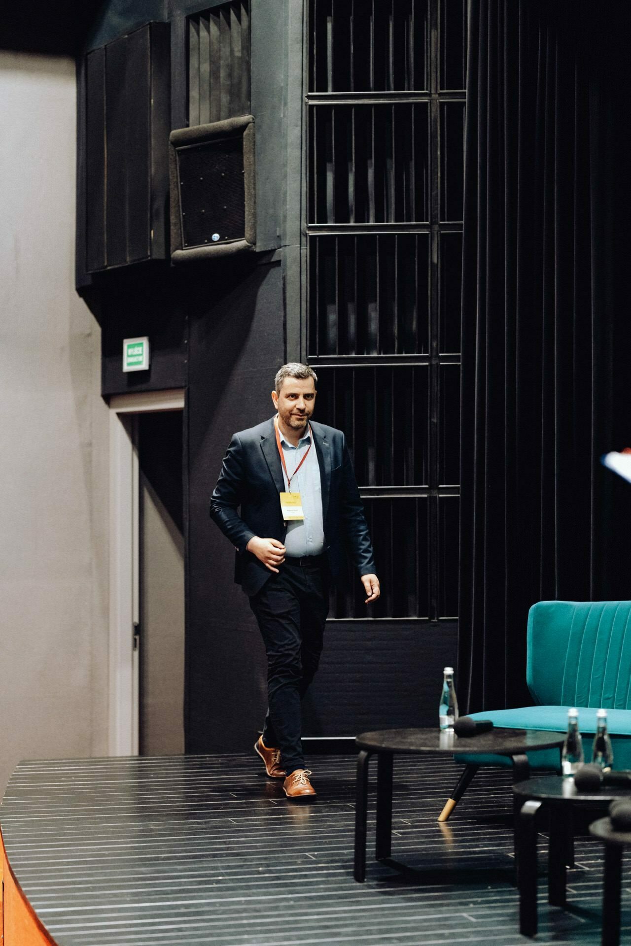 A man in a dark suit enters the scene with a black background. He has short hair and a beard, and wears a white shirt and brown shoes. On the stage, prepared for the photographer's event on, there is a green chair, a small table with water bottles and a briefcase. An emergency exit sign is visible in the background.   