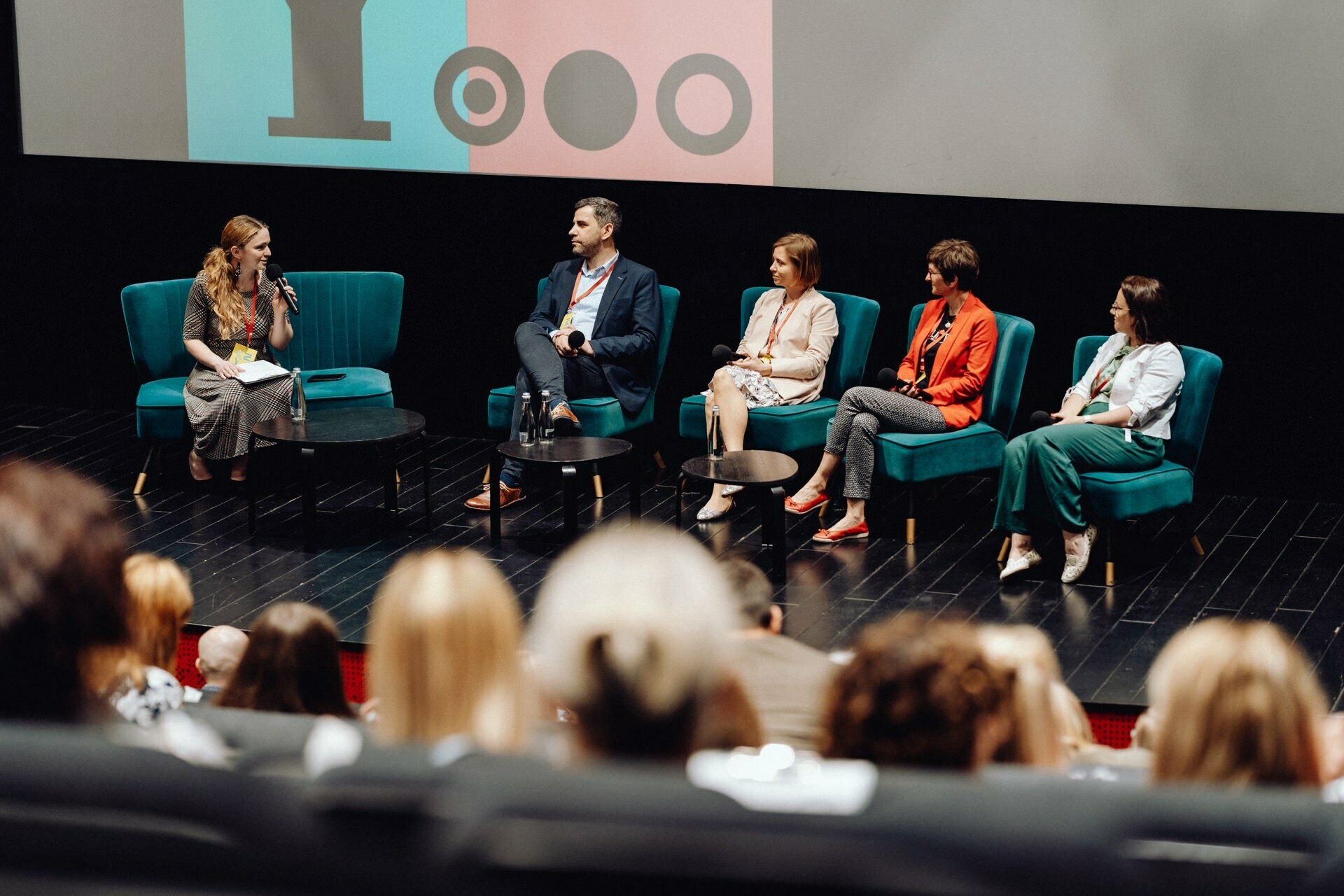 During the discussion, a panel of five sits on stage in turquoise chairs. The panelists are three women and two men, one of whom speaks into a microphone. The audience watches from their seats, with a large screen with graphics in the background. For those who want to capture such moments, consider hiring an event photographer who specializes in event photo coverage.   