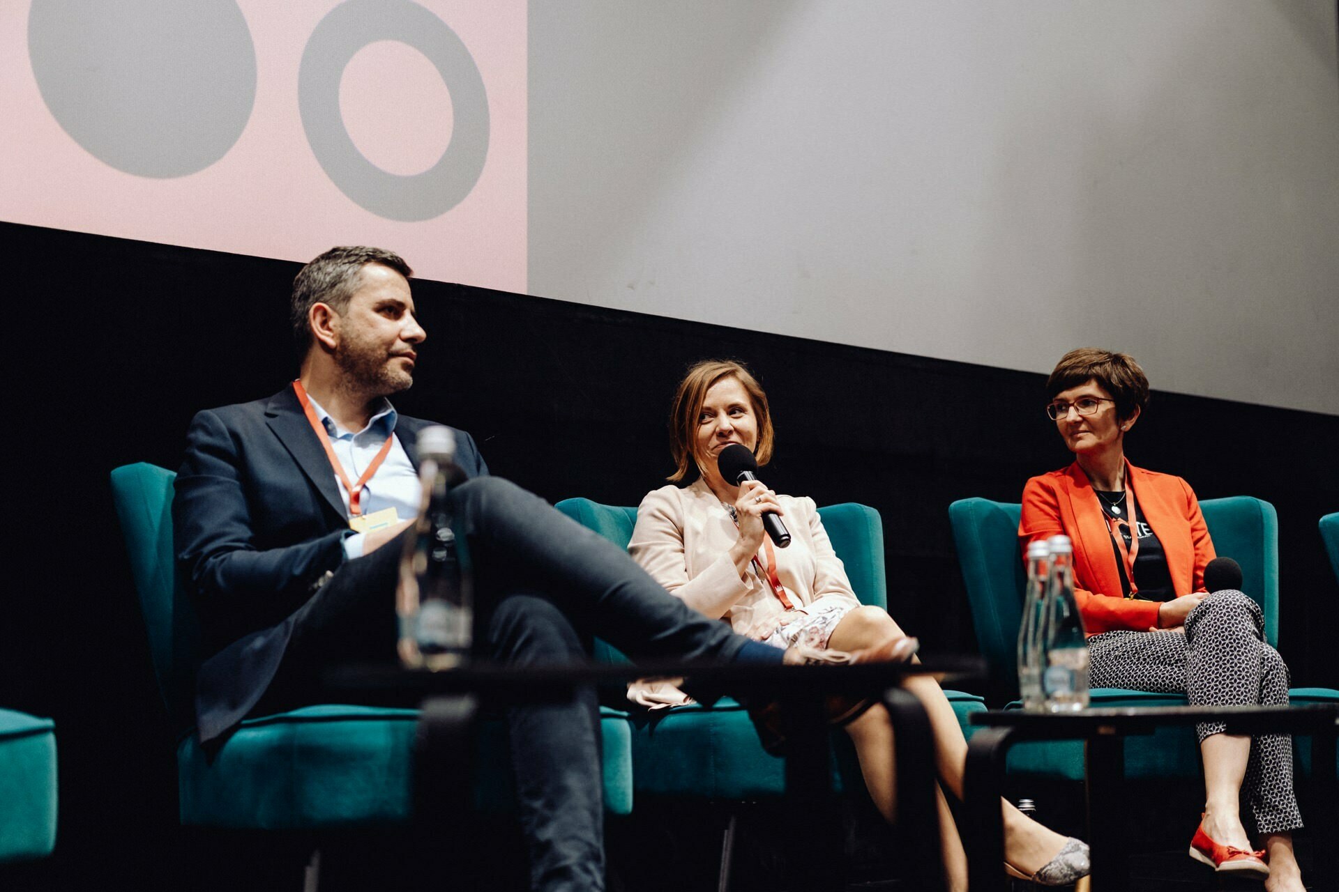 Three people sit on stage in turquoise chairs and take part in a panel discussion. The woman in the middle speaks into a microphone while the others listen intently. In front of them is a table with bottles of water, and in the background is a screen with wheels - perfect for photo coverage of the event.  