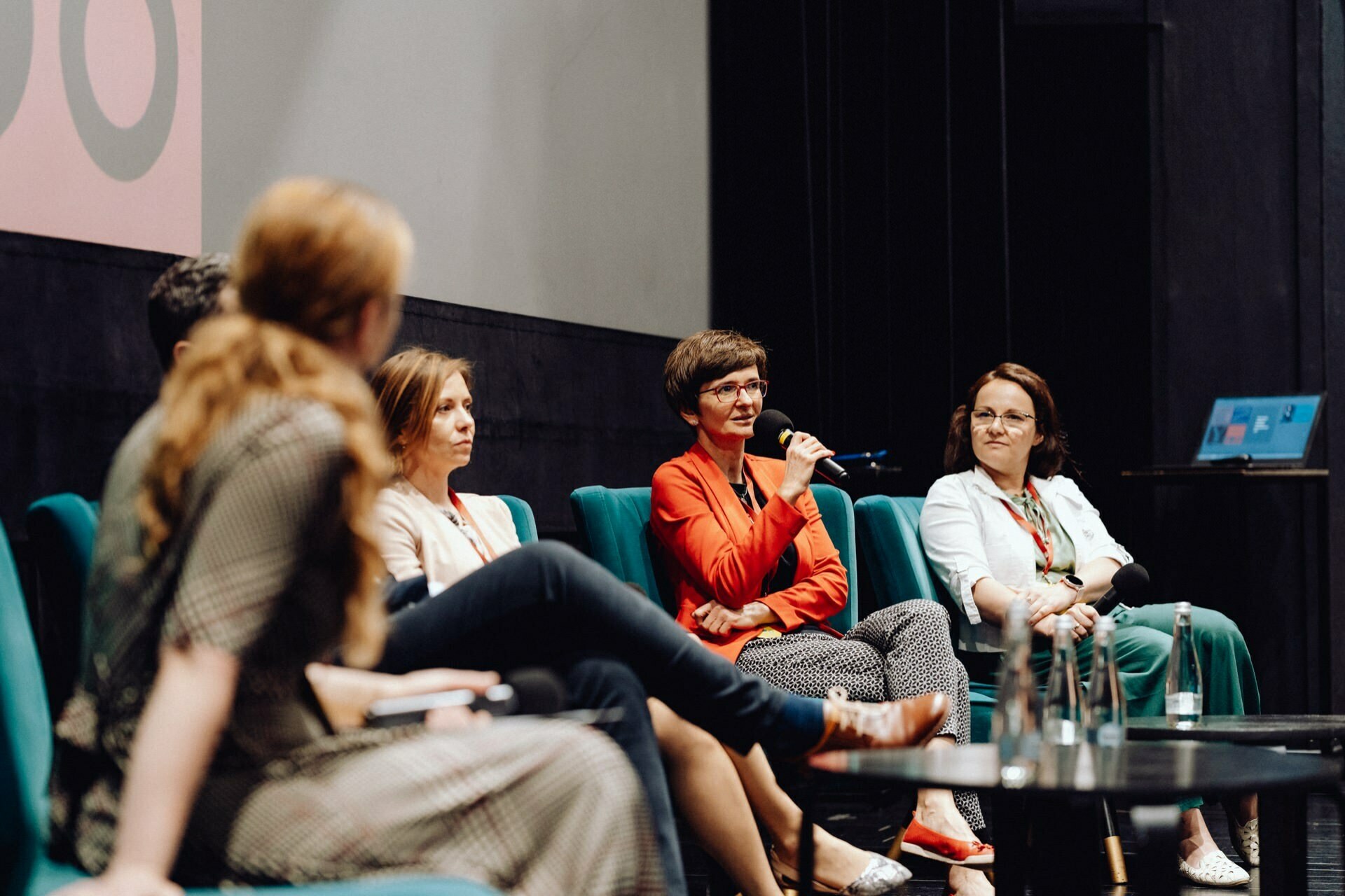 A group of five people sit on chairs on the stage and take part in a panel discussion. One person speaks into the microphone, while the others listen intently. Photos from the event beautifully show them lined up in an arch, with a table and water bottles in front of them.  