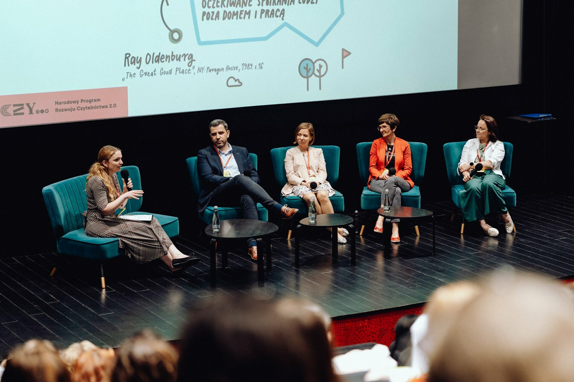 Five people sit on the stage and take part in a panel discussion. Four of them sit on colorful chairs, and one person speaks into a microphone. Behind them, a presentation slide with text and graphics is projected on a screen. The audience is visible in the foreground, captured by an event photographer warszawa.   