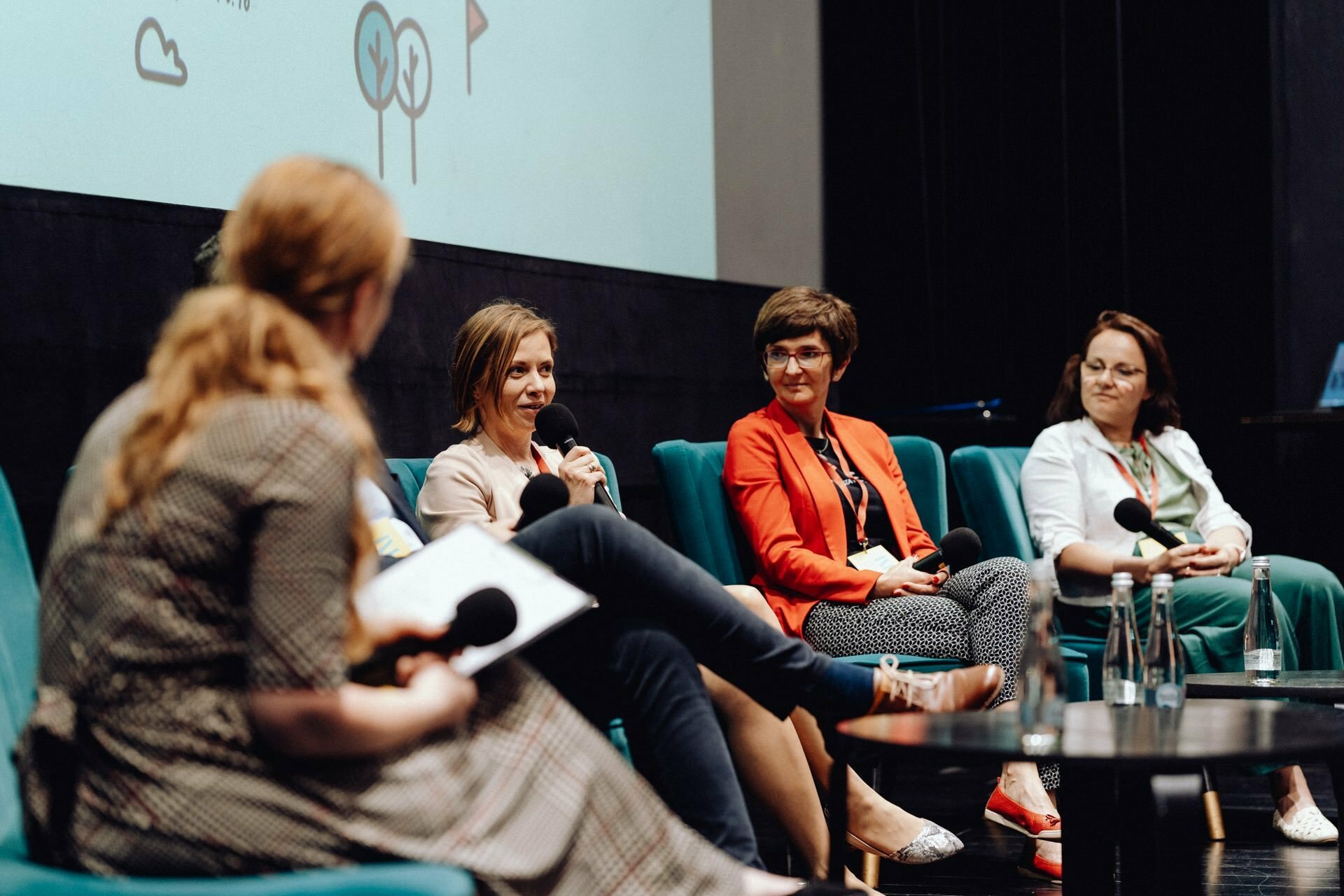 Four people are sitting in a panel discussion. A woman in a patterned dress is blurred in the foreground, holding a microphone. Three other women sit on blue chairs, facing the audience. Behind them is a white screen with a minimalist graphic of trees and clouds, captured by a photographer at the event.   