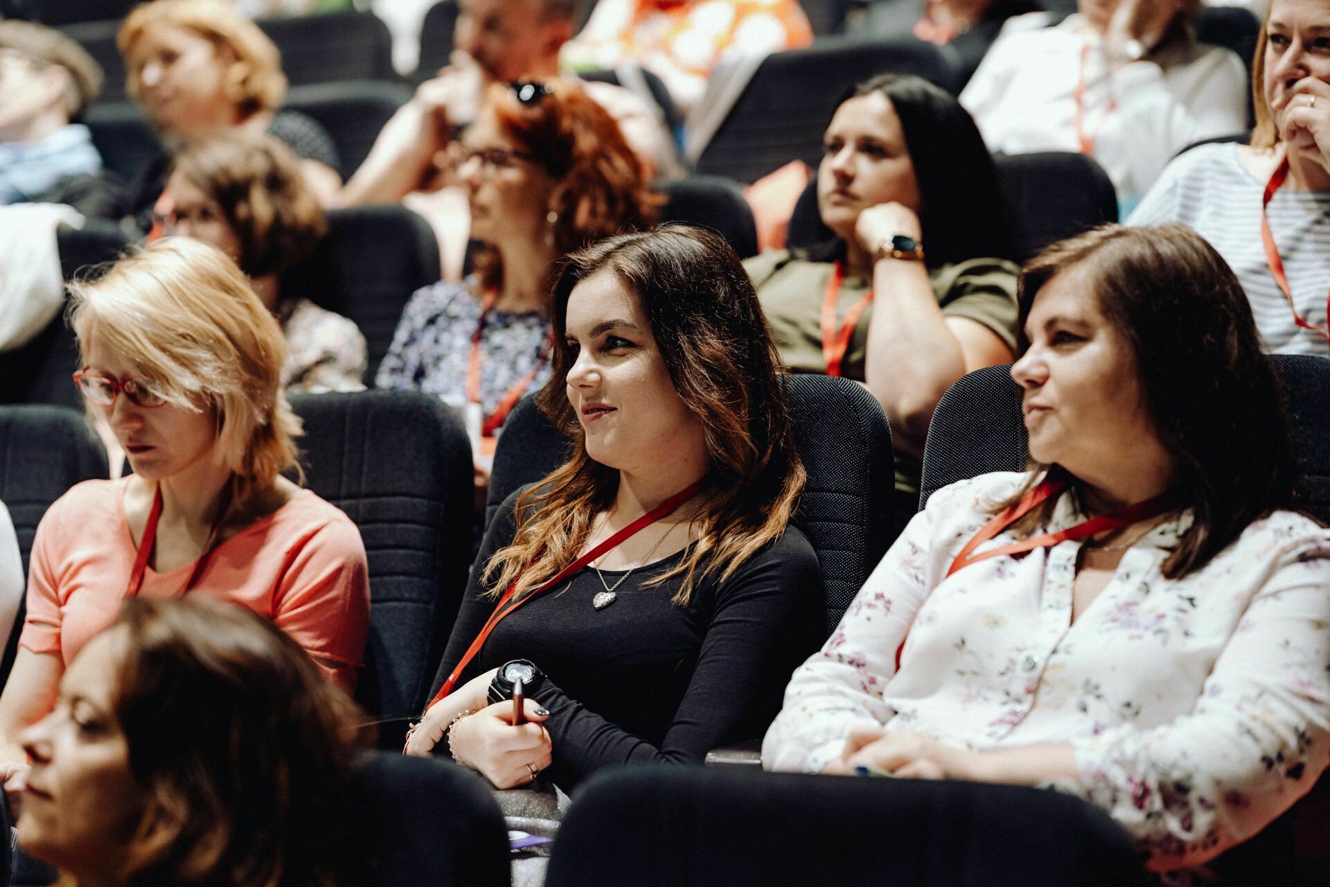 A diverse group of people sit in the auditorium, some focused and others engaged in conversation. They wear casual clothes and wear name badges on red lanyards, suggesting a seminar or conference. A photographer at the event captures every moment and creates a detailed photo report of the event.  