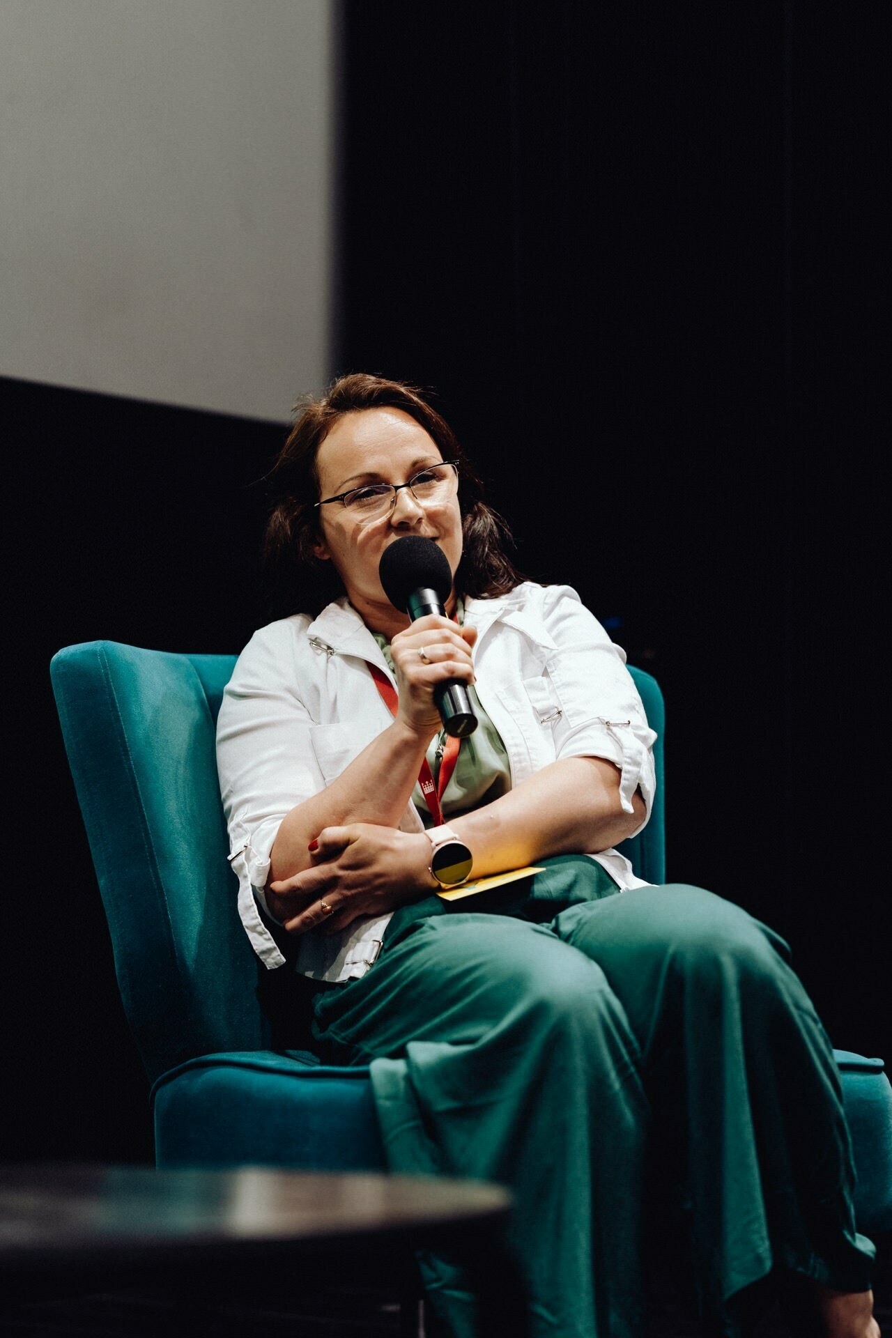 A person wearing glasses and a white shirt, sitting on a turquoise chair, holds a microphone and speaks. They have a thoughtful expression and are wearing green pants. The dark background contrasts with the speaker and the chair, highlighting them in the scene - perfect for a photo essay of the event.  