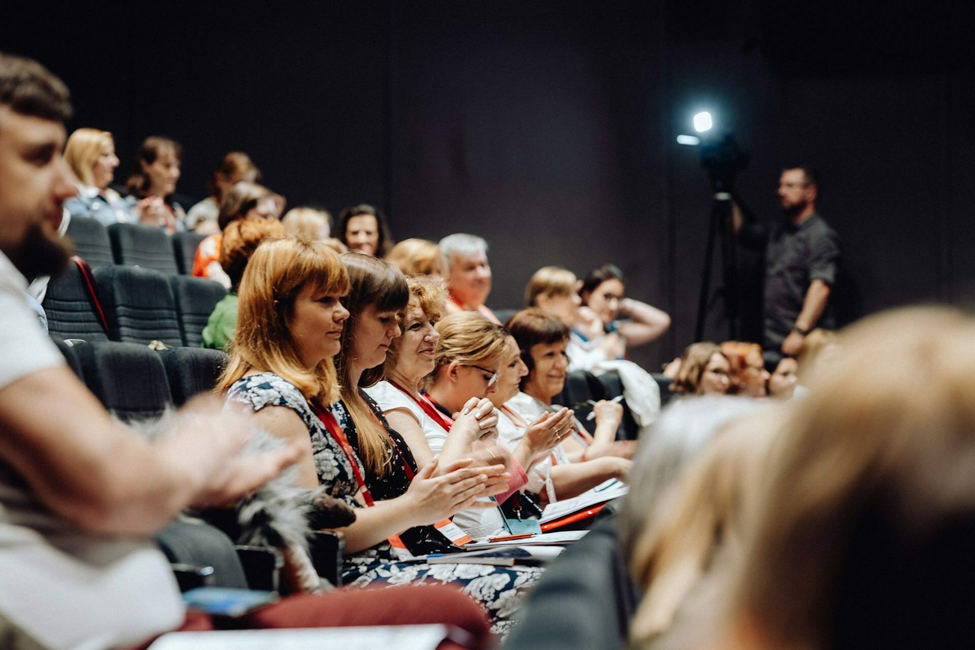 A diverse group of people sitting in the auditorium are intently watching the presentation. Some are clapping, others are taking notes. In the background, a person stands next to a set of cameras, presumably recording the event and providing a photo report of the event by a photographer from Warsaw.  