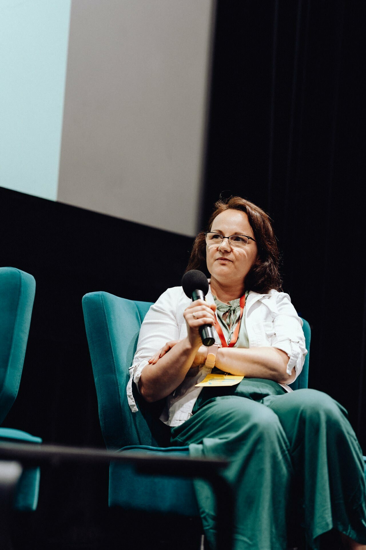 A person wearing a white shirt and green pants sits on a turquoise chair, holds a microphone and speaks. The background is dark, with a projection screen partially visible behind them. The scene looks as if it came from a photo report from an event or was captured by a photographer for an event.  