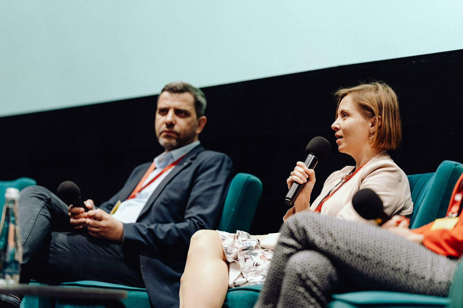 On stage, two people sit and speak into microphones. The person on the left is holding the microphone and looking ahead, while the person on the right is speaking, holding the microphone close to his mouth. Both are wearing conference badges - the perfect event photo by an event photographer.  