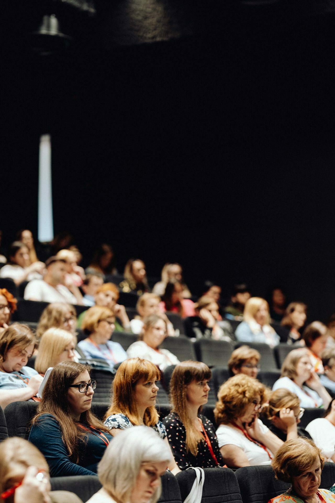 A large group of people sitting in an auditorium and carefully watching a presentation or event. The audience is diverse, being of different genders and ages. Captured by event photographer Warsaw, the seating areas are arranged in rows and the background is mostly dark, highlighting the participants.  