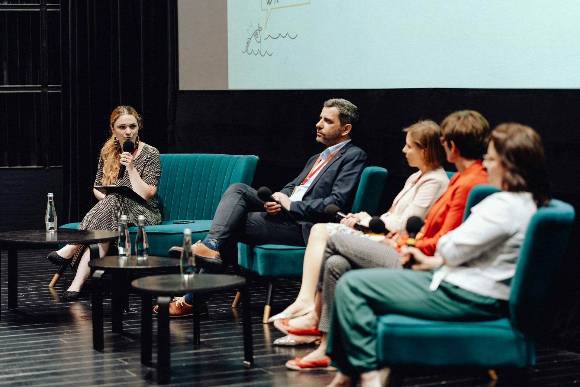 During the discussion, a panel of five sits on stage in turquoise chairs. Everyone is holding a microphone and busy talking. A woman on the left leads the discussion, and an event photographer Warsaw captures the moment. The background is a large, partially visible screen.   