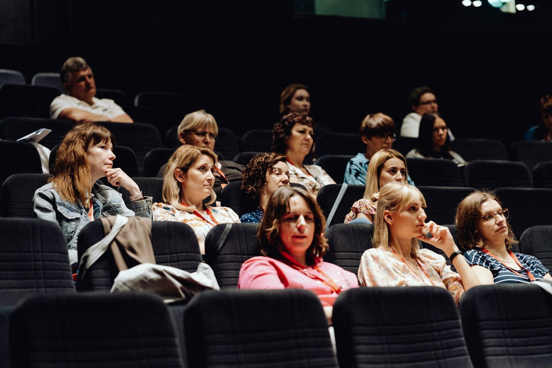 A group of people sit in rows in a dimly lit room and watch or listen intently to a presentation. They look engaged, some with thoughtful faces, others taking notes, which is beautifully captured by the event photographer warszawa. 