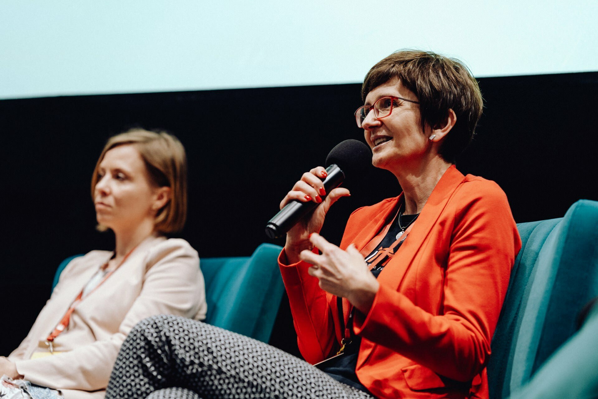 Two women are sitting on the stage. The woman on the right, wearing glasses and a red jacket, speaks into a microphone. The other woman, in a light-colored jacket, listens intently. This captivating event photo shows the two women deeply involved in a panel discussion or event.   