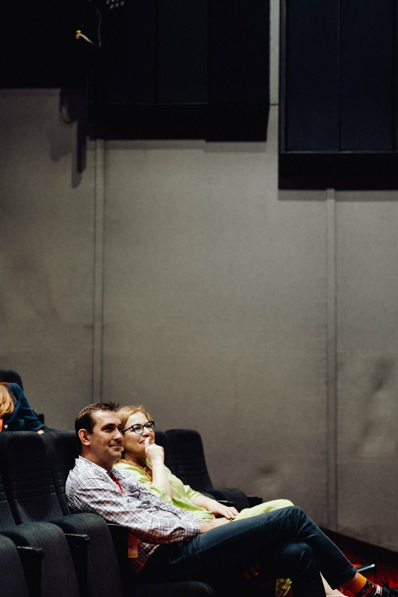 A man and a woman are sitting in a dark theater watching something intently. The man in the checkered shirt and the woman in the yellow top are sitting close to each other, both looking forward with relaxed and contented faces - a perfect moment captured by the photographer for the event. 