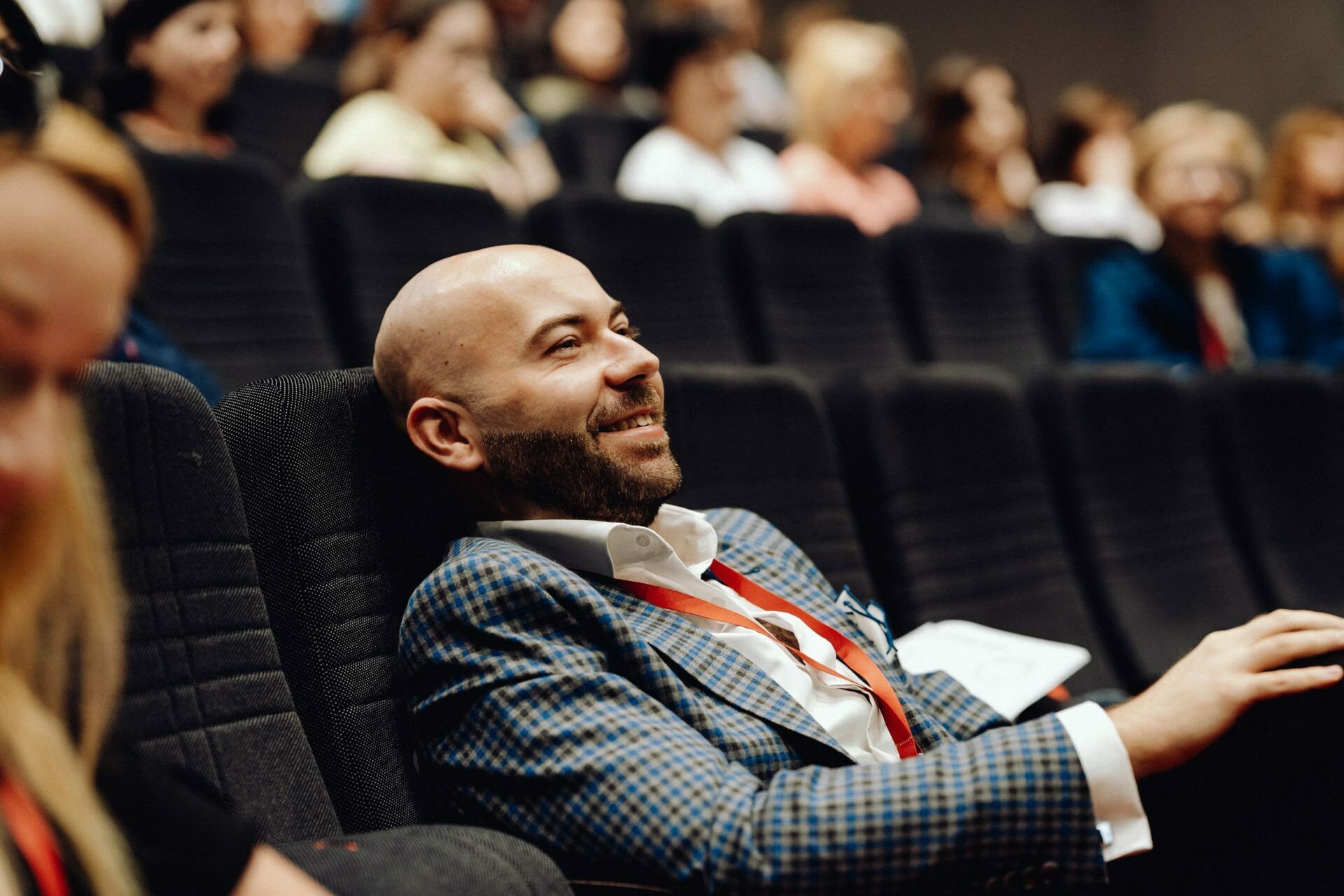 A bald bearded man in a checkered suit and red lanyard smiles and sits in a theater-style chair among a group of other seated people. Attention is focused on the man, while other people in the background are a bit out of focus, which was beautifully captured by a photographer from Warsaw who specializes in event photojournalism. 