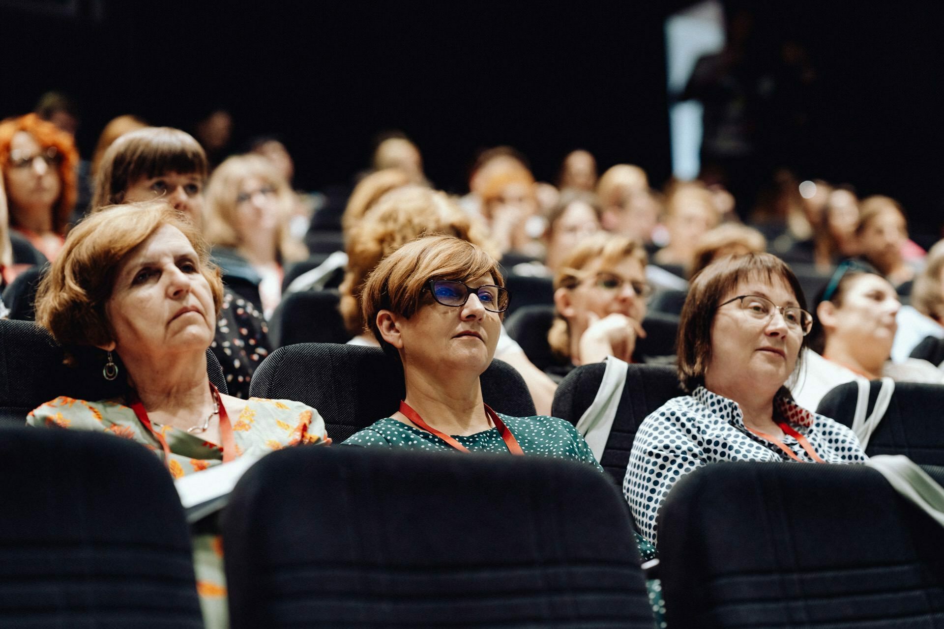 A group of people, mostly women, are sitting in a darkened room and carefully watching a presentation. Some have notebooks and lanyards, indicating that this may be a conference or seminar. A photographer from Warsaw captured the moment when the background is filled with other seated attendees.  