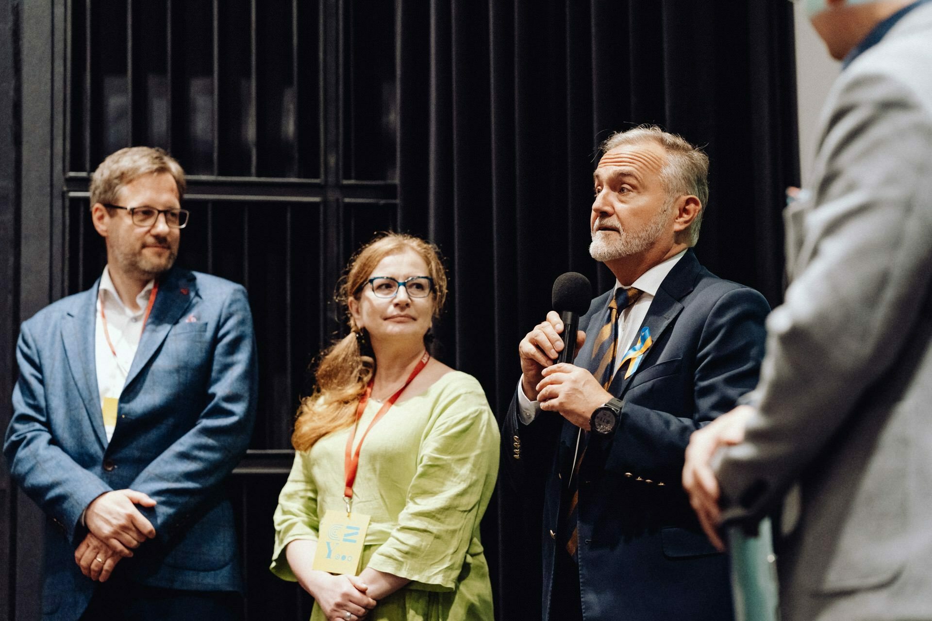 A group of four people gather in conversation. A man in a suit speaks into a microphone, while a woman and another man, both with lanyards, listen intently. The event photographer warsaw captured the moment when the fourth person, only partially visible, is standing to the right.  