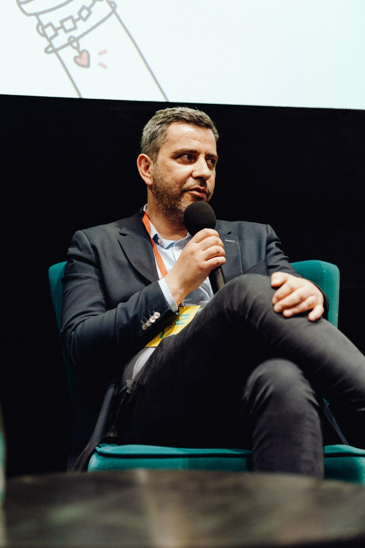 A man in a suit, sitting on a turquoise chair, is speaking into a microphone, which is perfectly captured by an event photographer Warsaw. He has short hair and beard, and his legs are crossed, giving the impression of focus. The dark background covers part of the screen displaying an image or presentation, perfect for a photo essay of the event.  