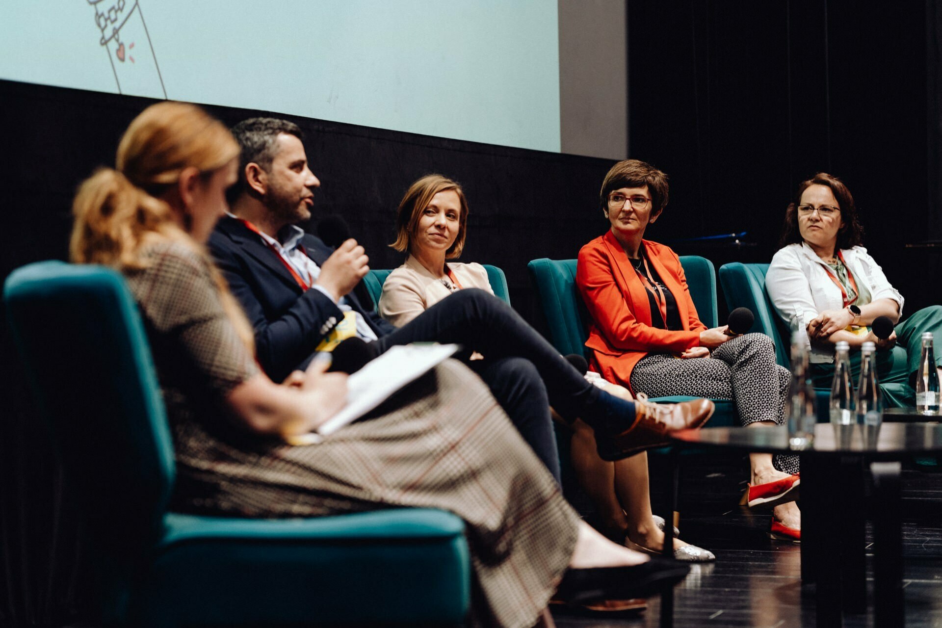 A five-member panel sits on stage immersed in discussion. Three women and one man sit in a row of turquoise chairs, while one woman, with her back turned to the camera, appears to be moderating the conversation. This scene, captured by a photographer at the event, depicts the lively dialogues against the backdrop of a large screen.  