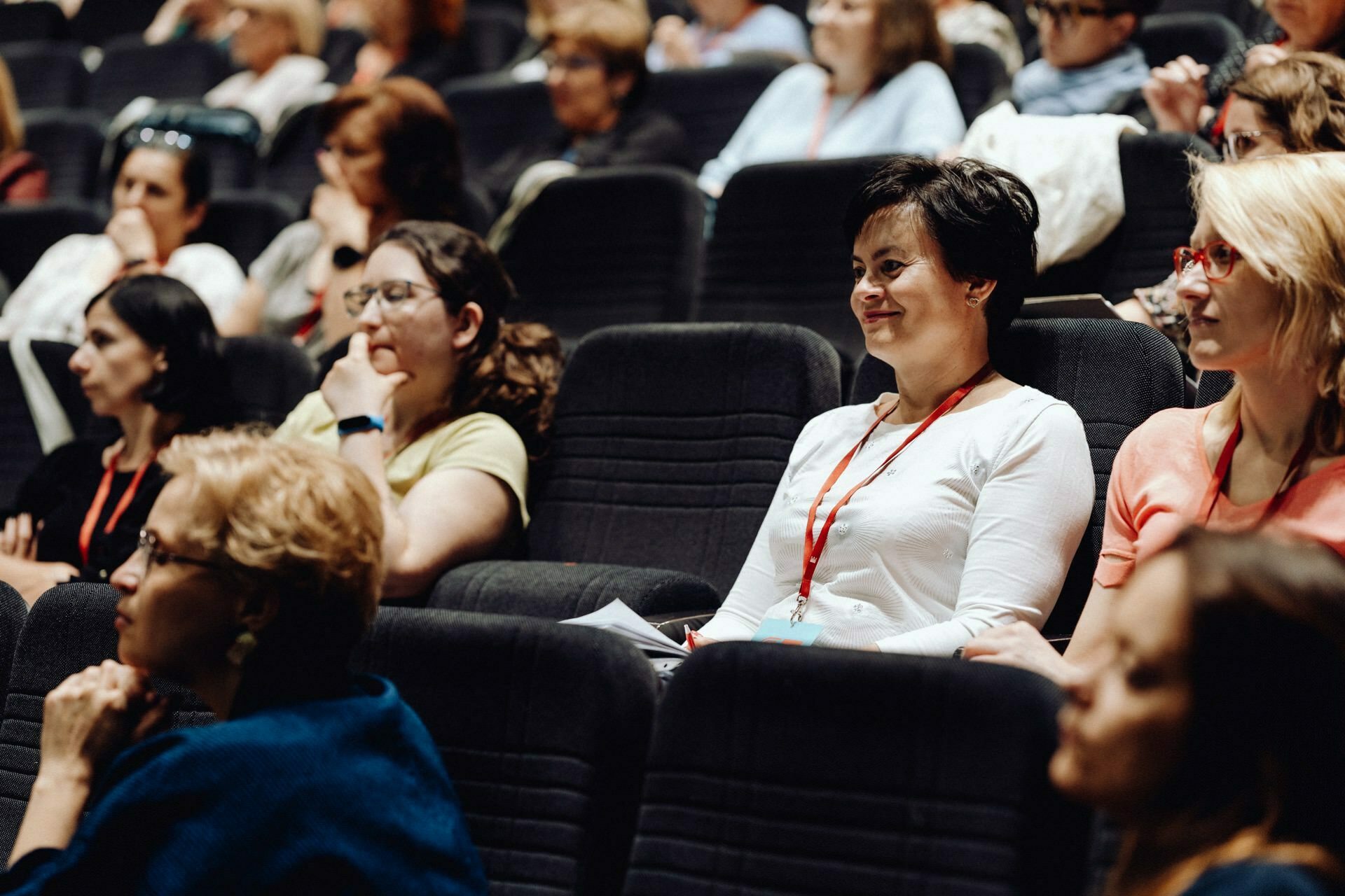 During the event, a diverse group of people, mostly women, sit in the auditorium. Many wear lanyards with name badges. Some listen intently, others take notes or look thoughtful. Black seats are lined up in rows - perfect for a photo essay of the event by a photographer from Warsaw.   