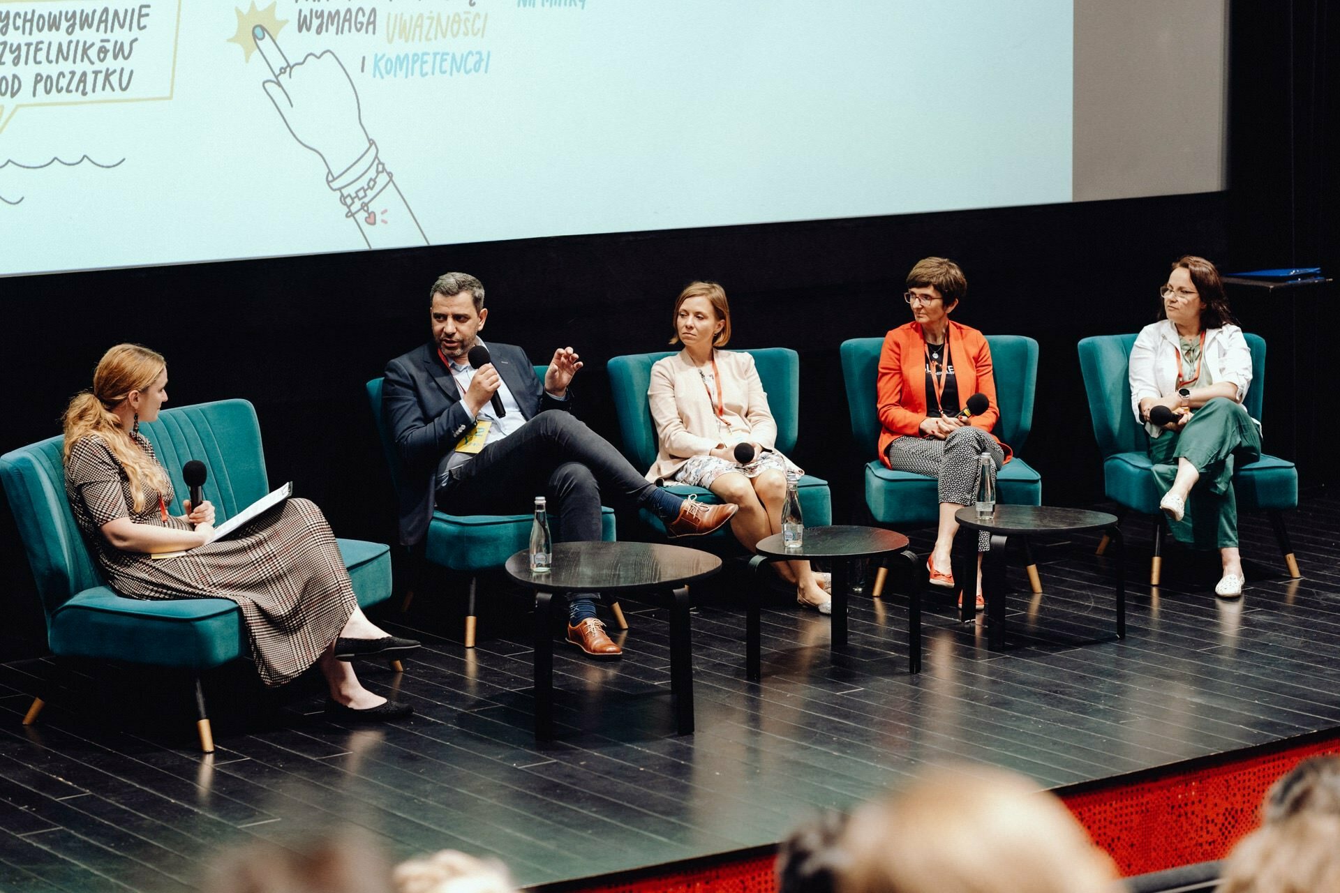 Five people sit on stage immersed in a panel discussion. Four of them are holding microphones. Three women and two men sit on green chairs with a black floor and a projector screen displaying illustrations. The audience, captured by a photographer at the event, is partially visible in the foreground.   