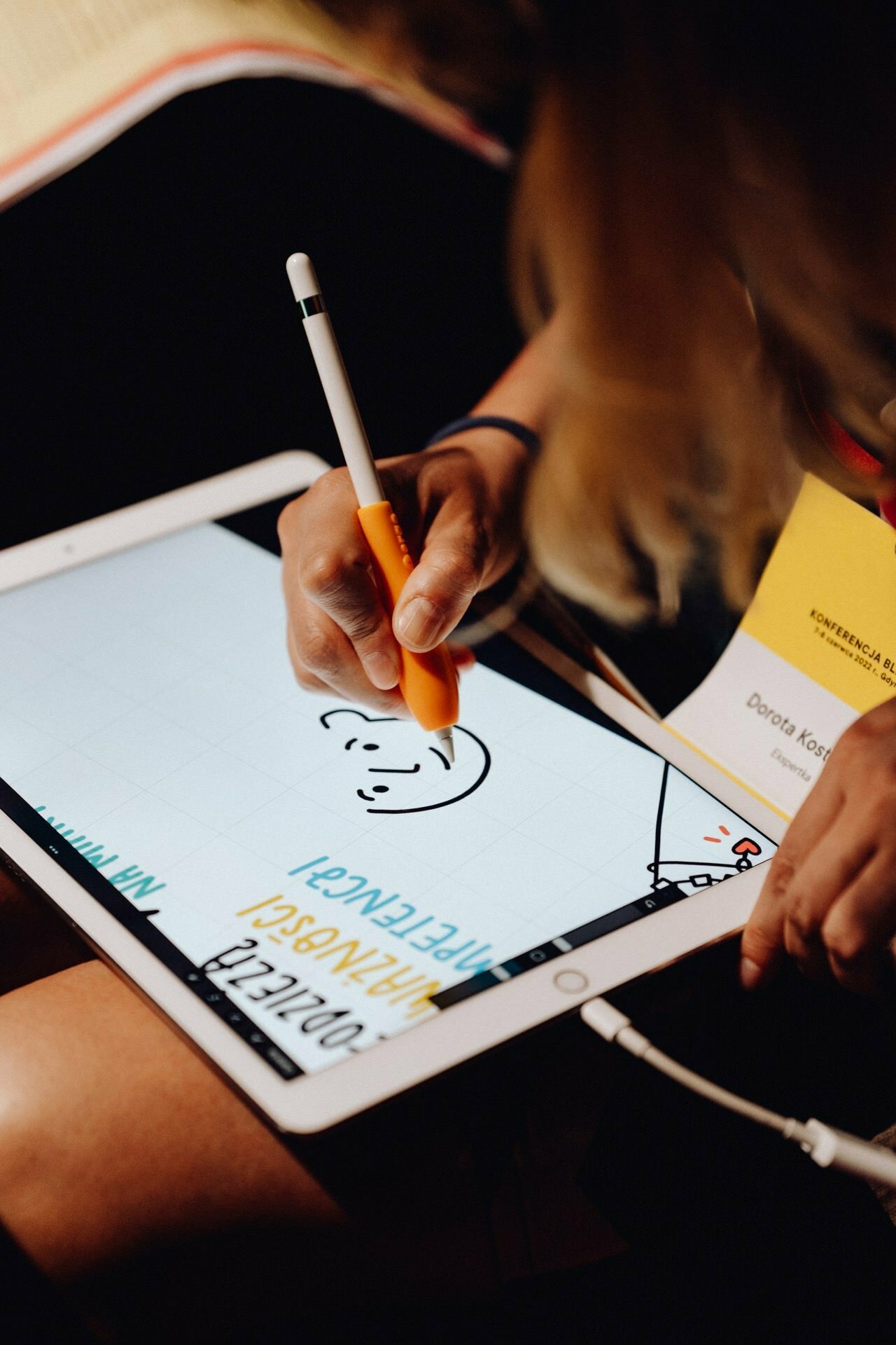 A person using a stylus to draw on a tablet. A drawing of a smiling face appears on the screen. A yellow conference badge with the name "Dorota Koto" is partially visible in the background, and the person is holding an orange pen. Nearby, an event photographer Warsaw captures the moment.   
